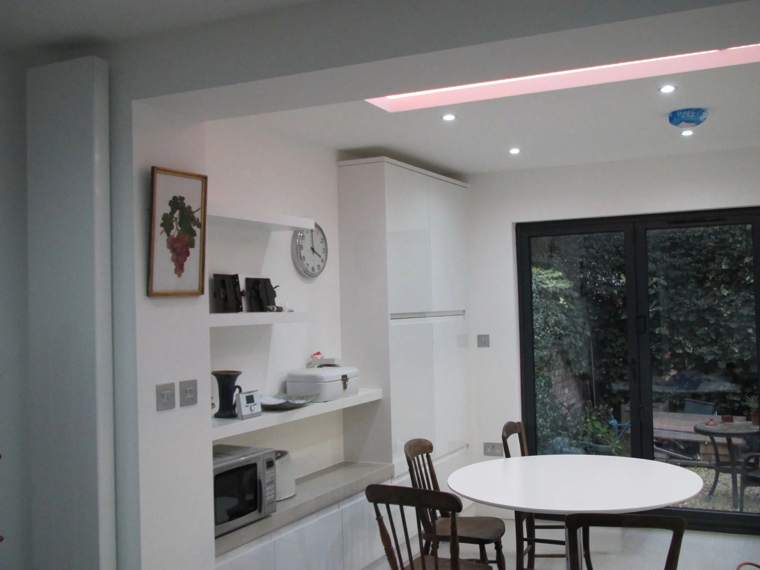 Kitchen and dining area with floating shelves, tall white units and a round dining table at a rear kitchen extension on Short Road, Chiswick, W4 by Robin Services.