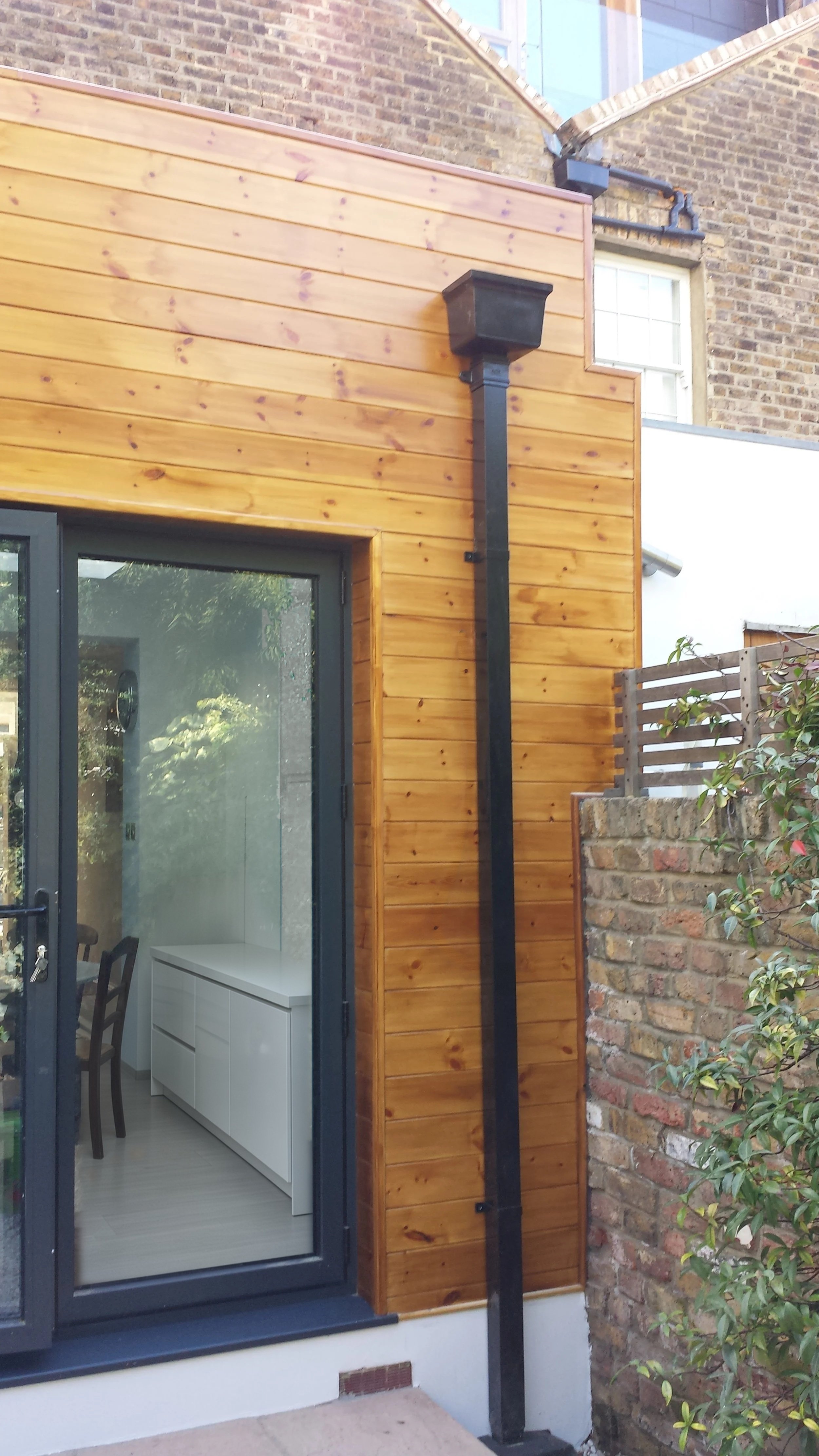 Pine-clad rear extension with a black drainpipe and a grey aluminium glazed door looking into a white gloss kitchen, at a rear kitchen extension on Short Road, Chiswick, W4 by Robin Services.
