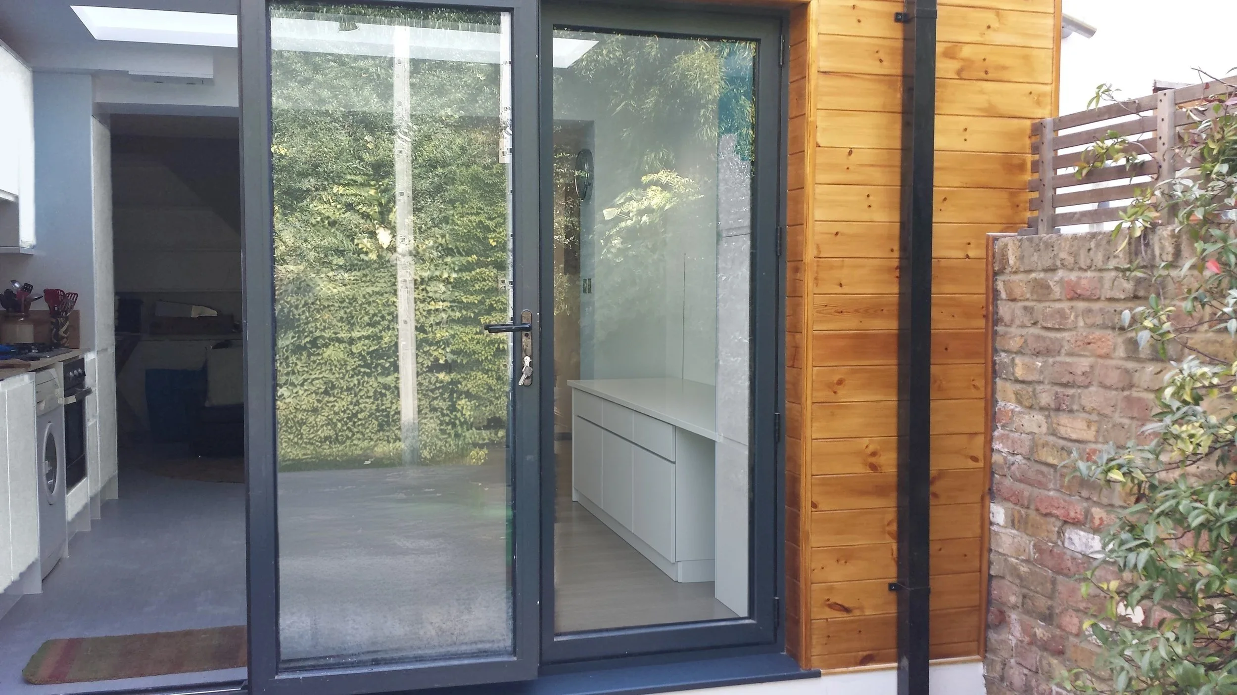 Closed grey aluminium sliding glass doors beside natural timber cladding, with a white kitchen and garden reflections in the glass, at a rear kitchen extension on Short Road, Chiswick, W4 by Robin Services.