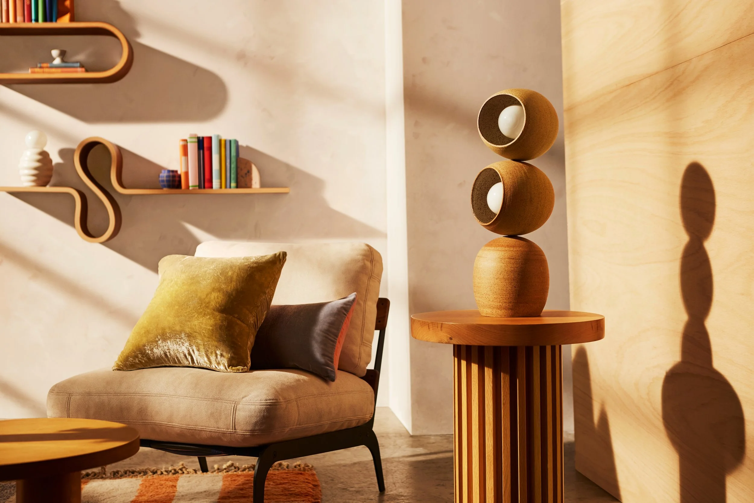 Interior of a living room featuring a beige sofa with pillows, suspended wooden shelves with books and decorative objects, and a wooden side table with stacked rounded vases casting shadows on the wall.