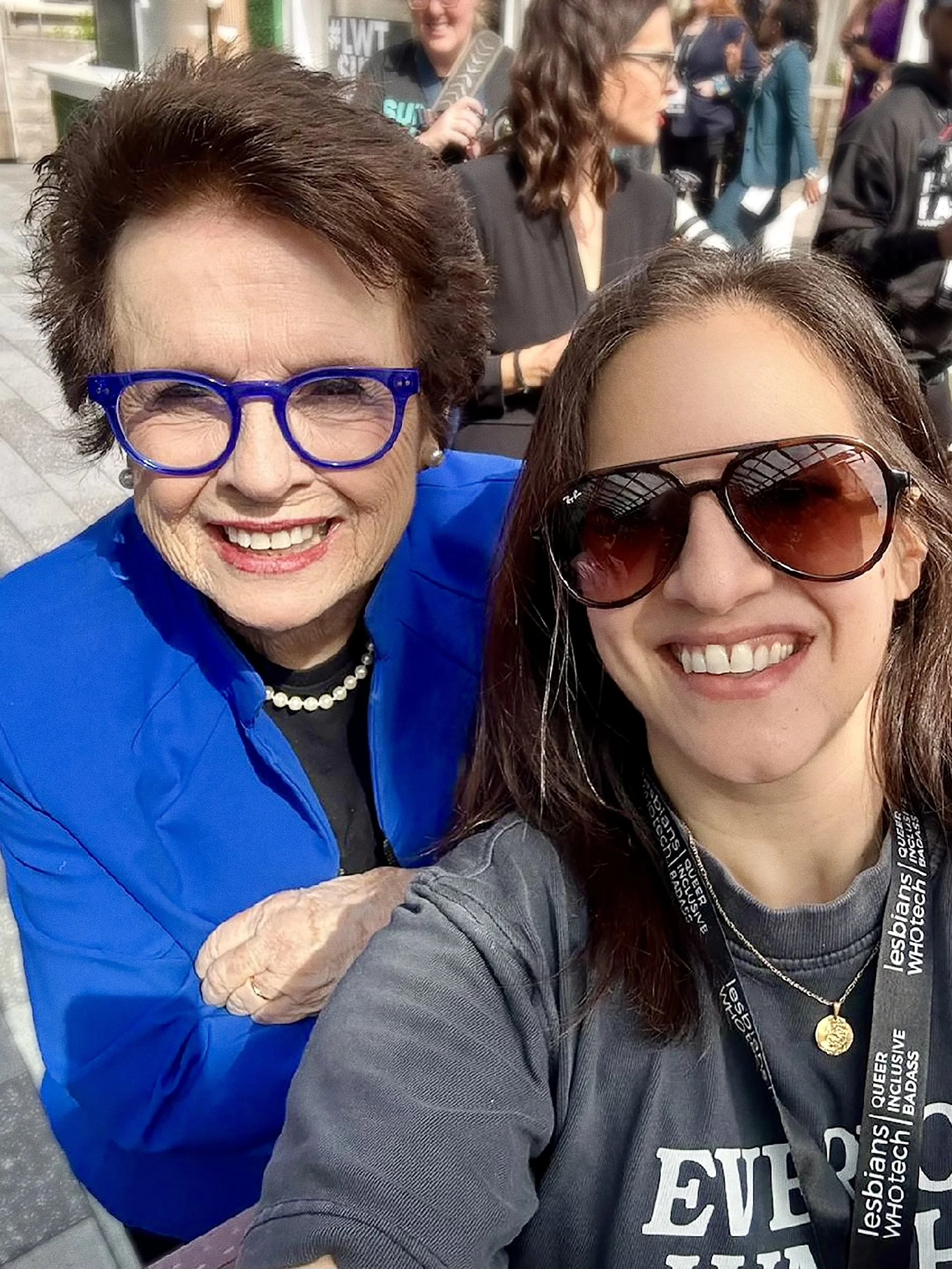 Two women smiling for a selfie at an outdoor event, with several other people in the background.