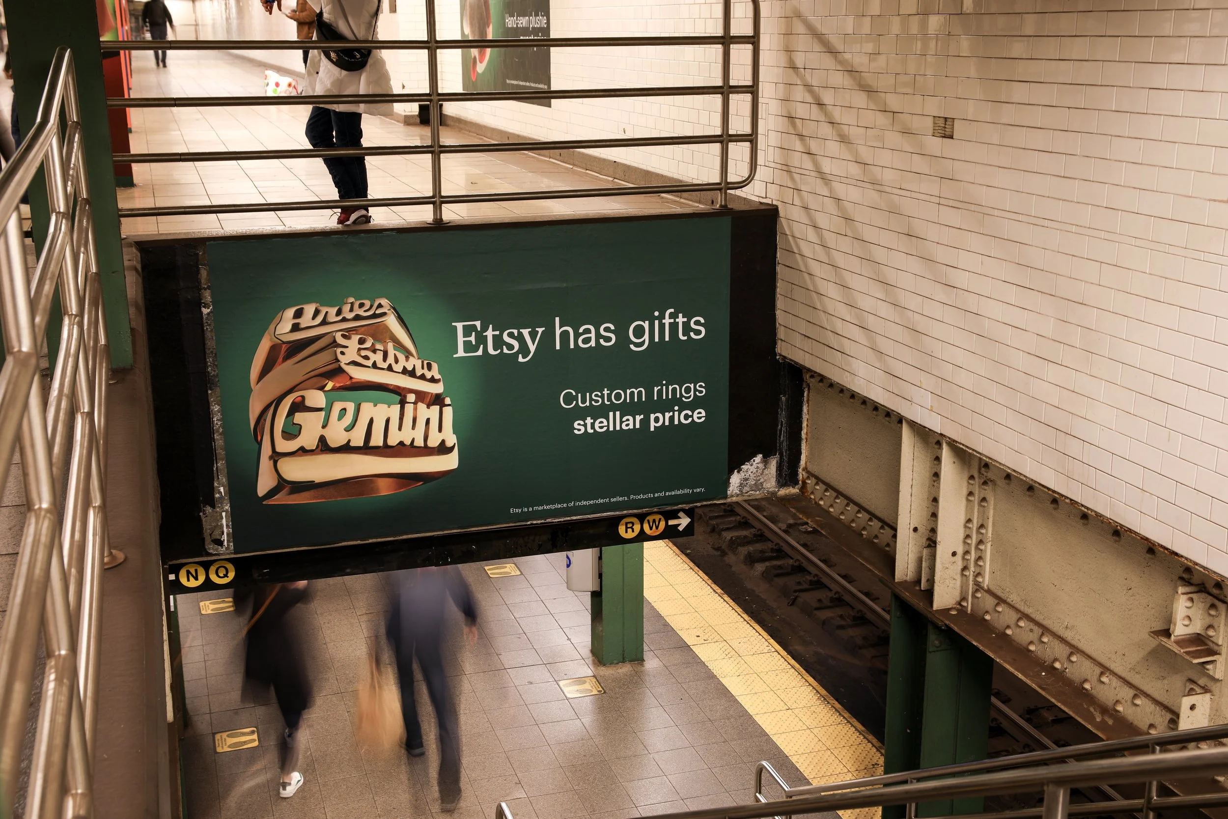 Subway station with an advertising sign for Etsy featuring a large graphic of custom rings and the text 'Etsy has gifts, Custom rings stellar price.' The platform shows blurred people walking, and the subway tracks are visible below. Metal railings are on the edges of the staircases.