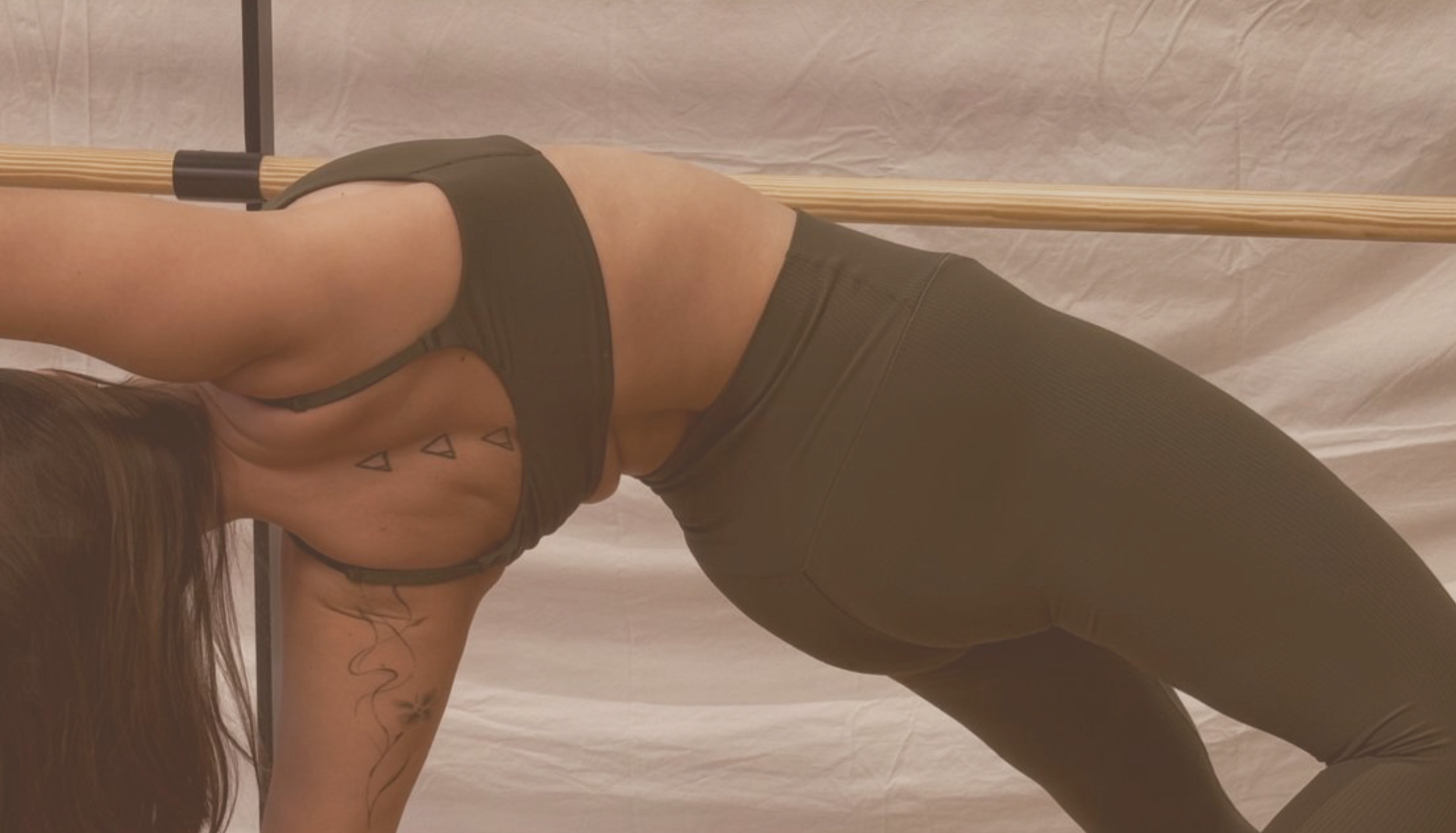 Instructora en postura de Barre, apoyada en manos y rodillas, con una barra de ballet detrás, vistiendo ropa deportiva de color oscuro.