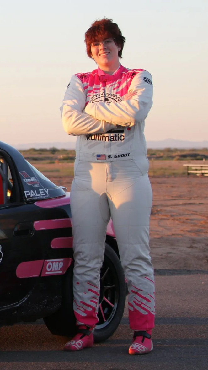 Person in racing suit standing next to race car in an open desert landscape during sunset.