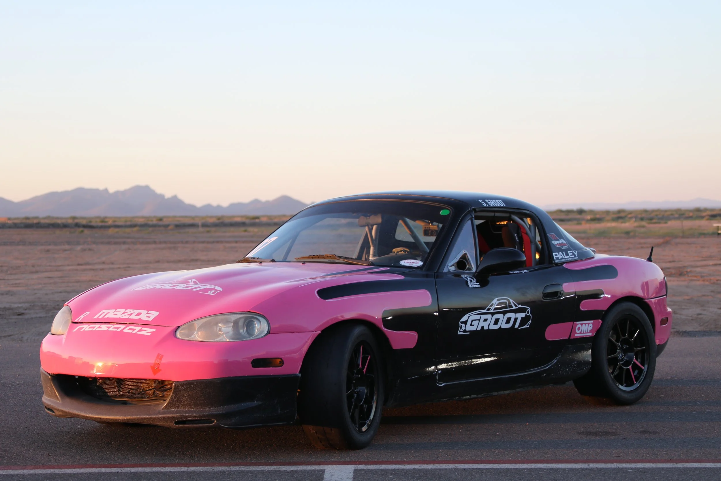 A pink and black race car with various sponsor logos, including Mazda and Groots, parked on a desert-like surface with mountains in the background during sunset.