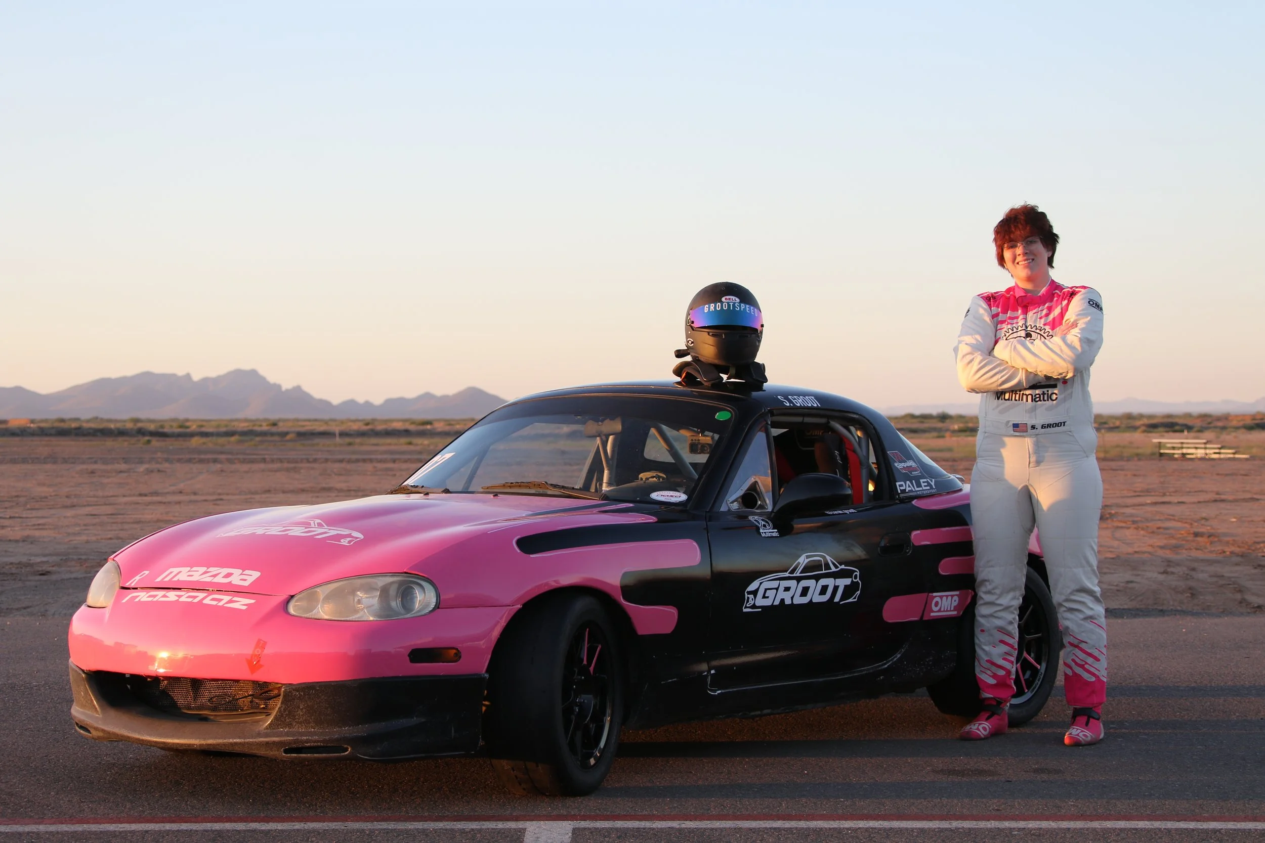 A race car driver in a white racing suit with pink accents standing next to a pink and black Mazda race car with sponsor logos, in a desert landscape with mountains in the background at sunset.
