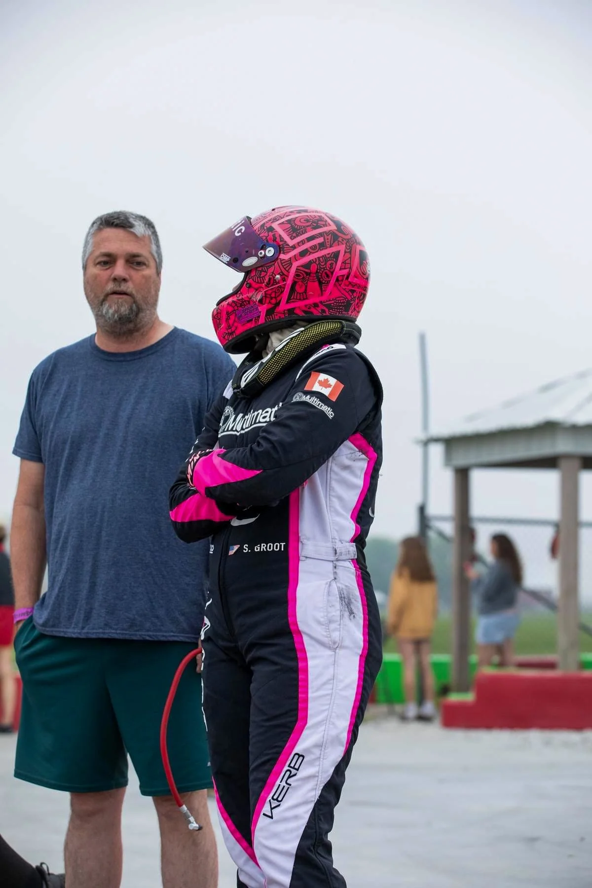 A man and a woman standing outdoors at a racing event. The woman is wearing a pink and black racing suit and helmet, with her arms crossed. The man is wearing a blue t-shirt and shorts, holding a racing helmet in his hand.