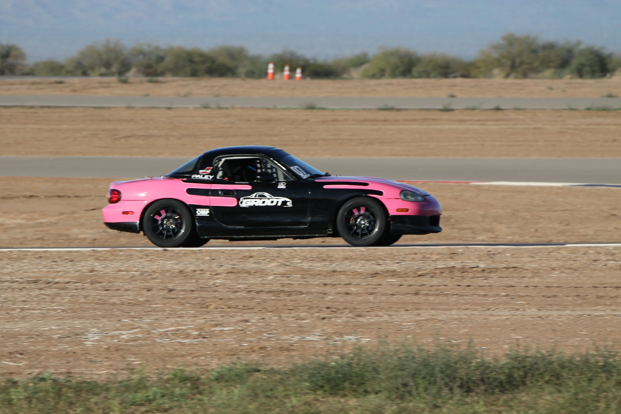 A pink and black race car driving on a race track in a desert landscape.