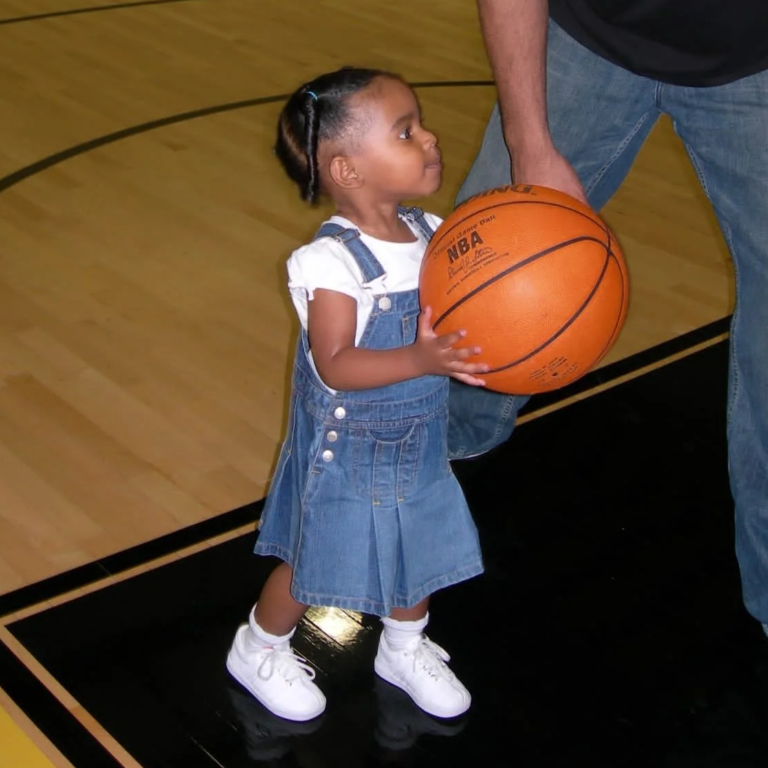 Young girl in denim dress and white sneakers holding a basketball on a basketball court.
