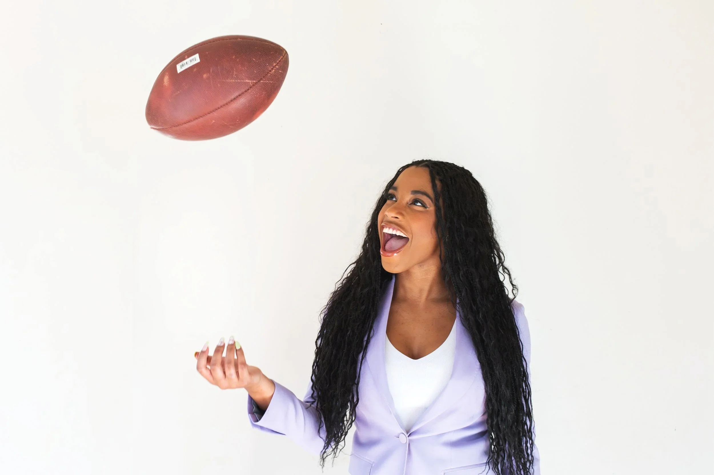 A woman with long curly black hair, wearing a light purple blazer and white top, smiling and looking towards a football floating in the air.