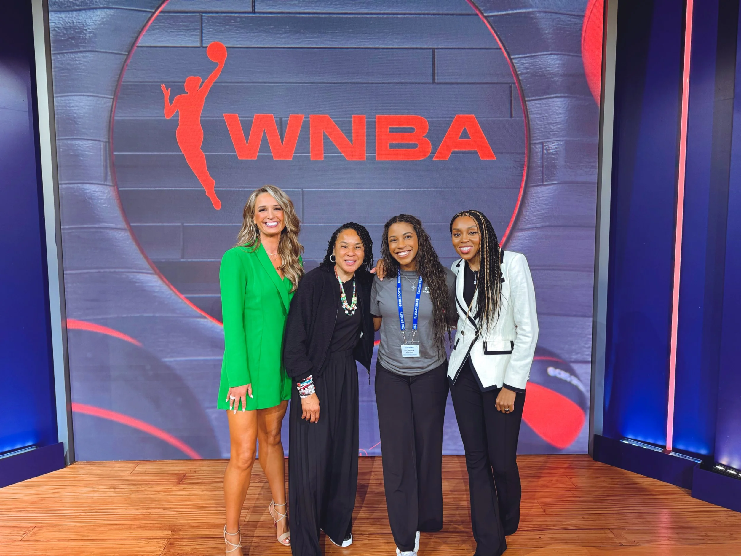 Four women standing together in front of a Women’s National Basketball Association (WNBA) logo, smiling for a photo.