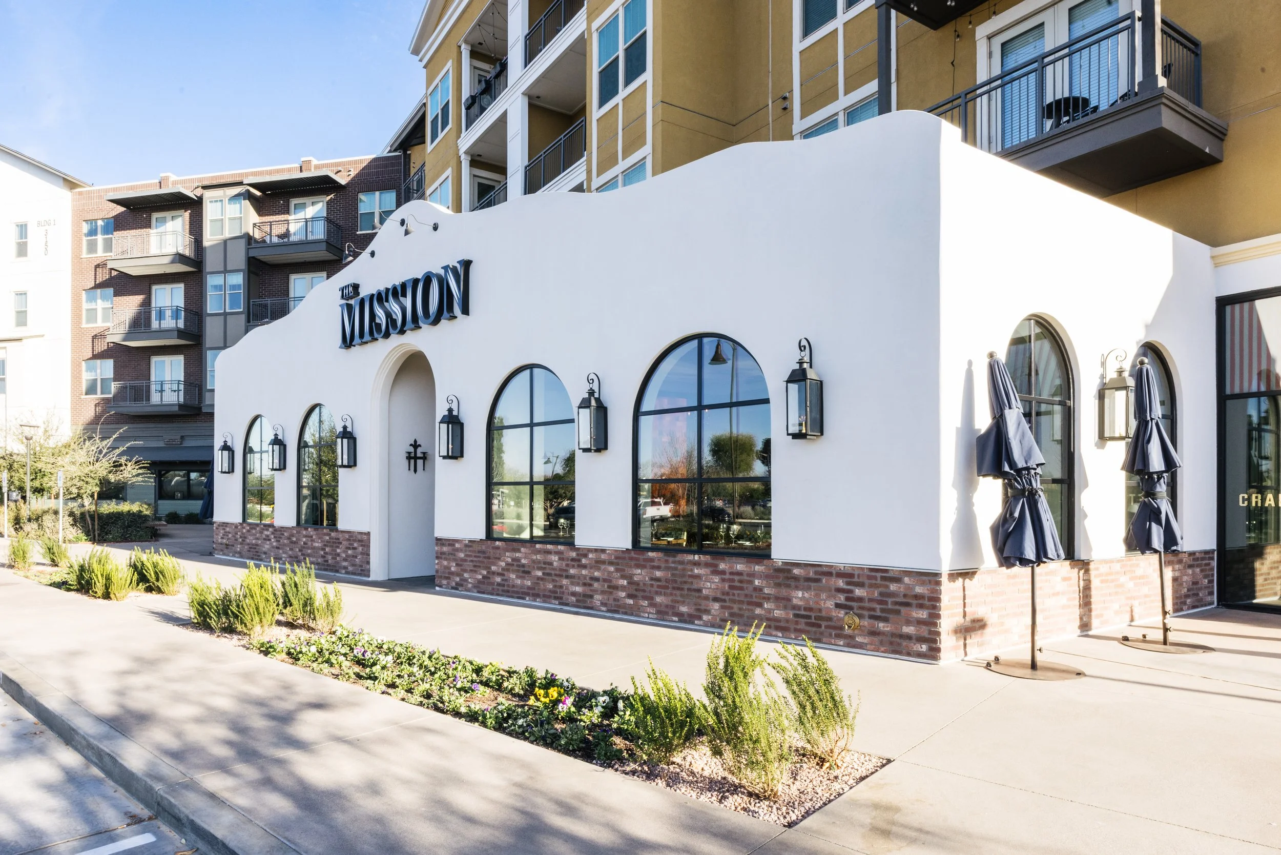 A modern residential building with balconies and a commercial storefront named 'The Mission' in an urban area during dusk, with a woman standing on the sidewalk and a few people walking nearby.