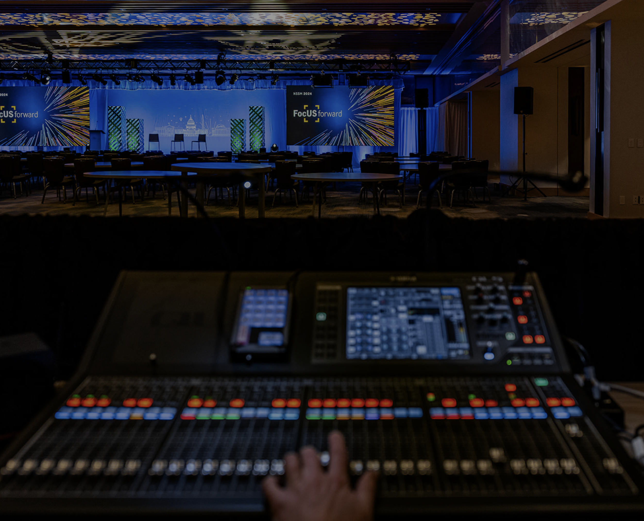 An auditorium or conference hall with rows of chairs and tables set for a formal event, facing a stage with a patterned backdrop and two large screens displaying presentation slides.