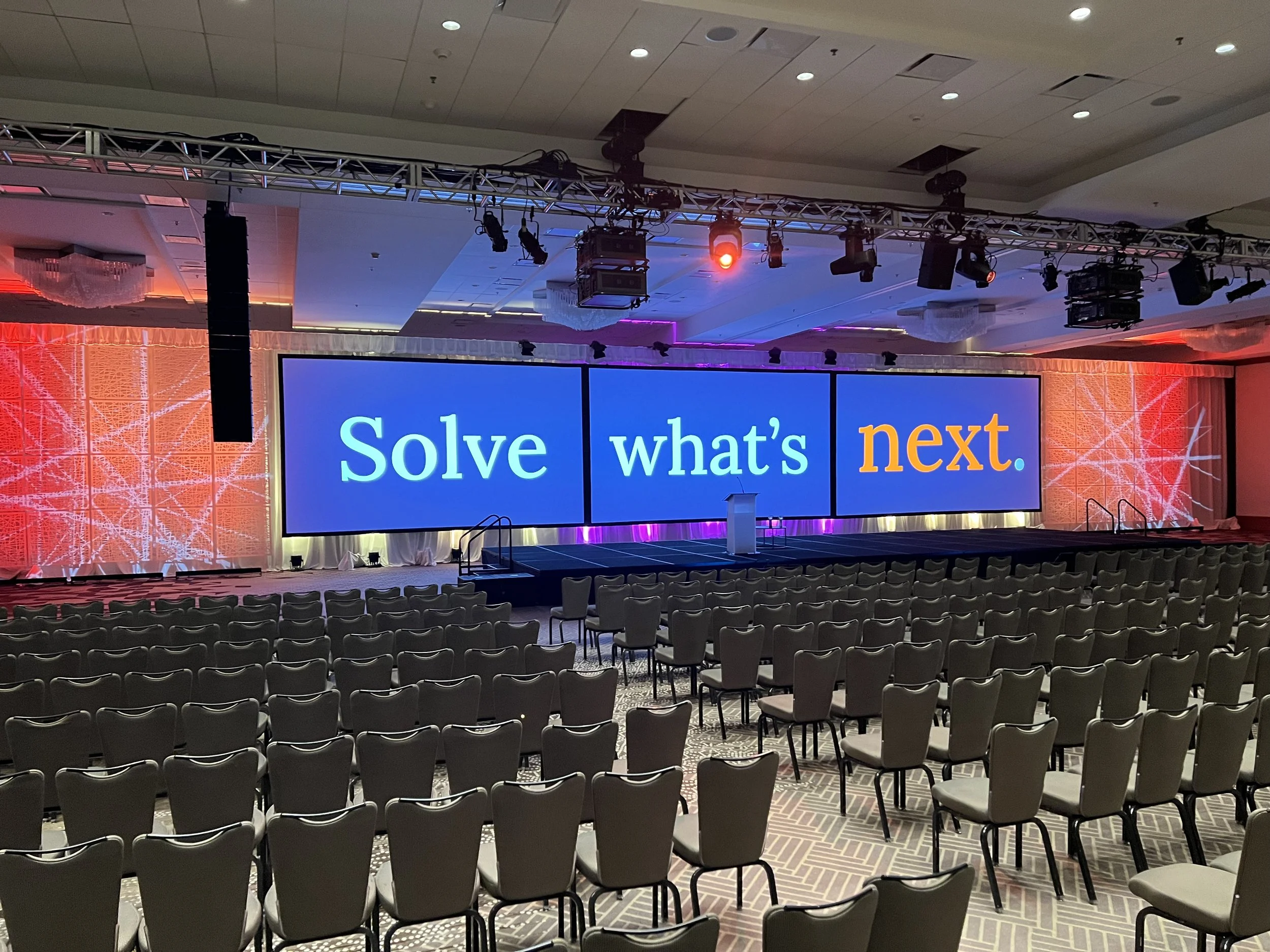 Empty conference room with rows of chairs facing a large digital screen displaying the phrase, 'Solve what's next.'