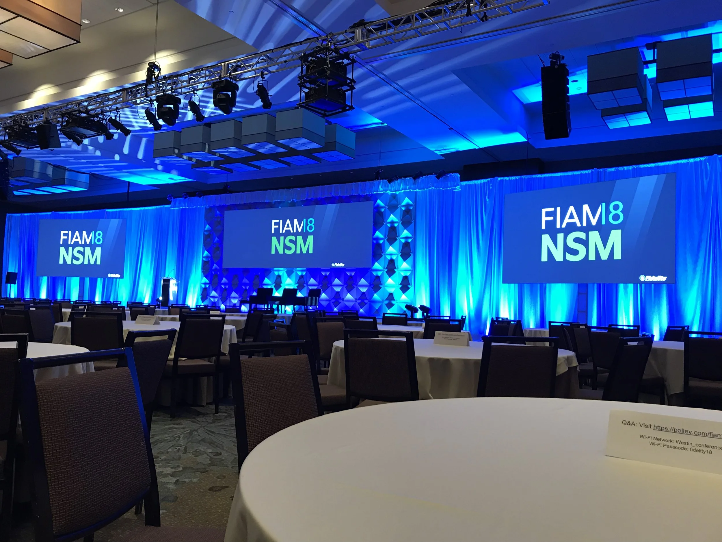 Conference room with round tables and chairs, set up for an event with a large stage and two big screens