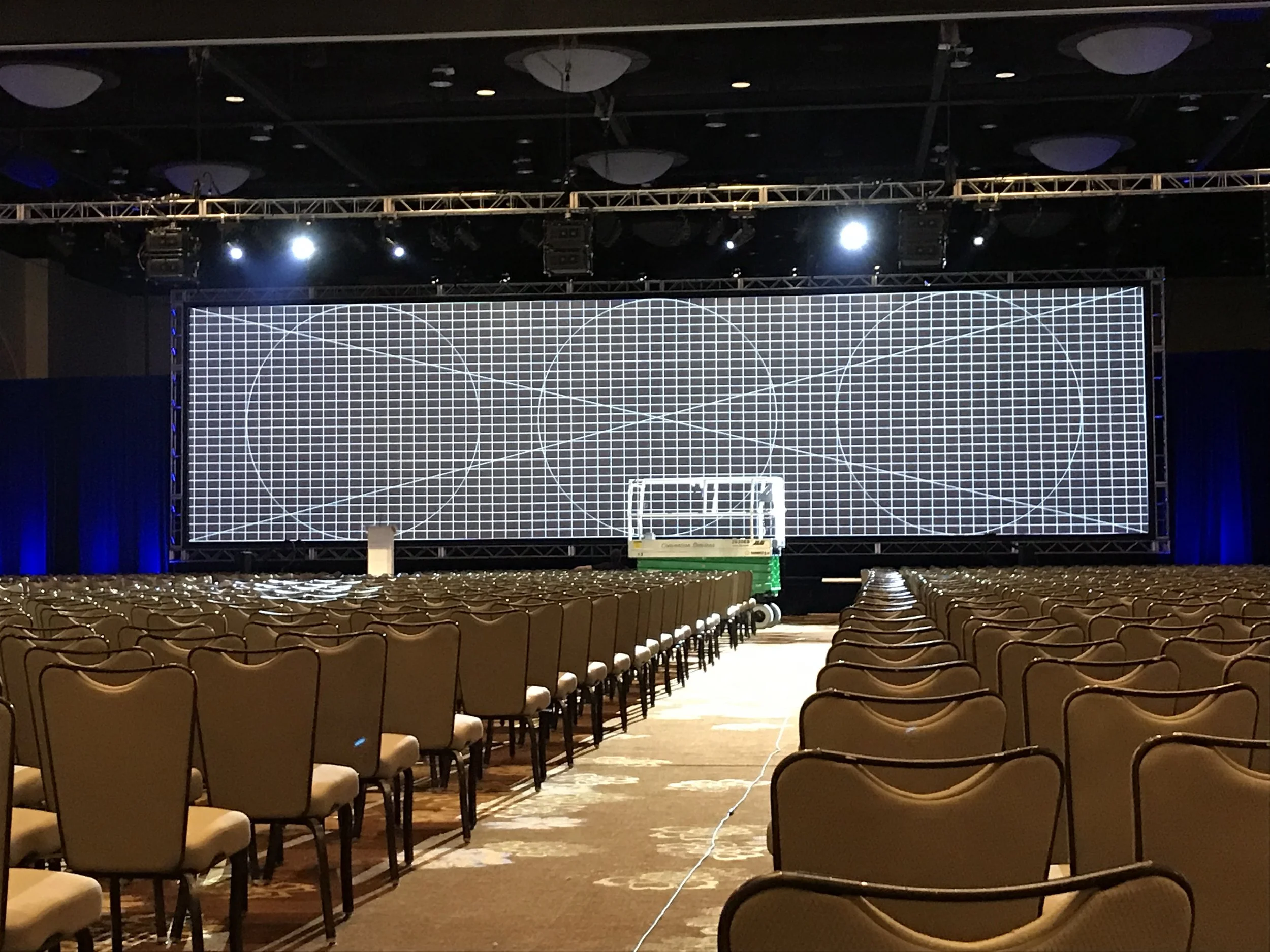 Empty conference or event hall with rows of chairs facing a large digital screen with grid lines and geometric designs, stage lighting overhead.