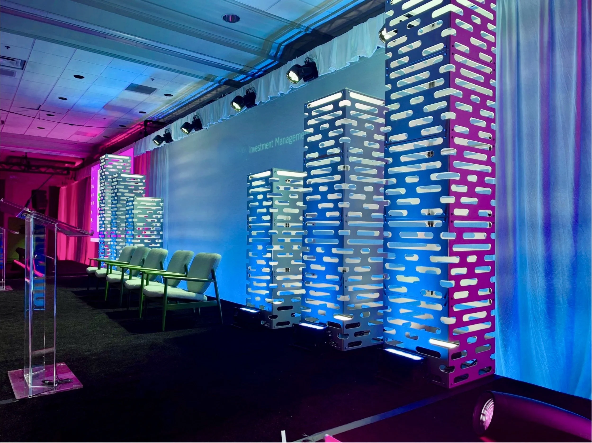 Stage setup with illuminated modular columns, row of chairs, and a lectern, prepare for a presentation or panel discussion at a conference, with colorful lighting and a large screen in the background.