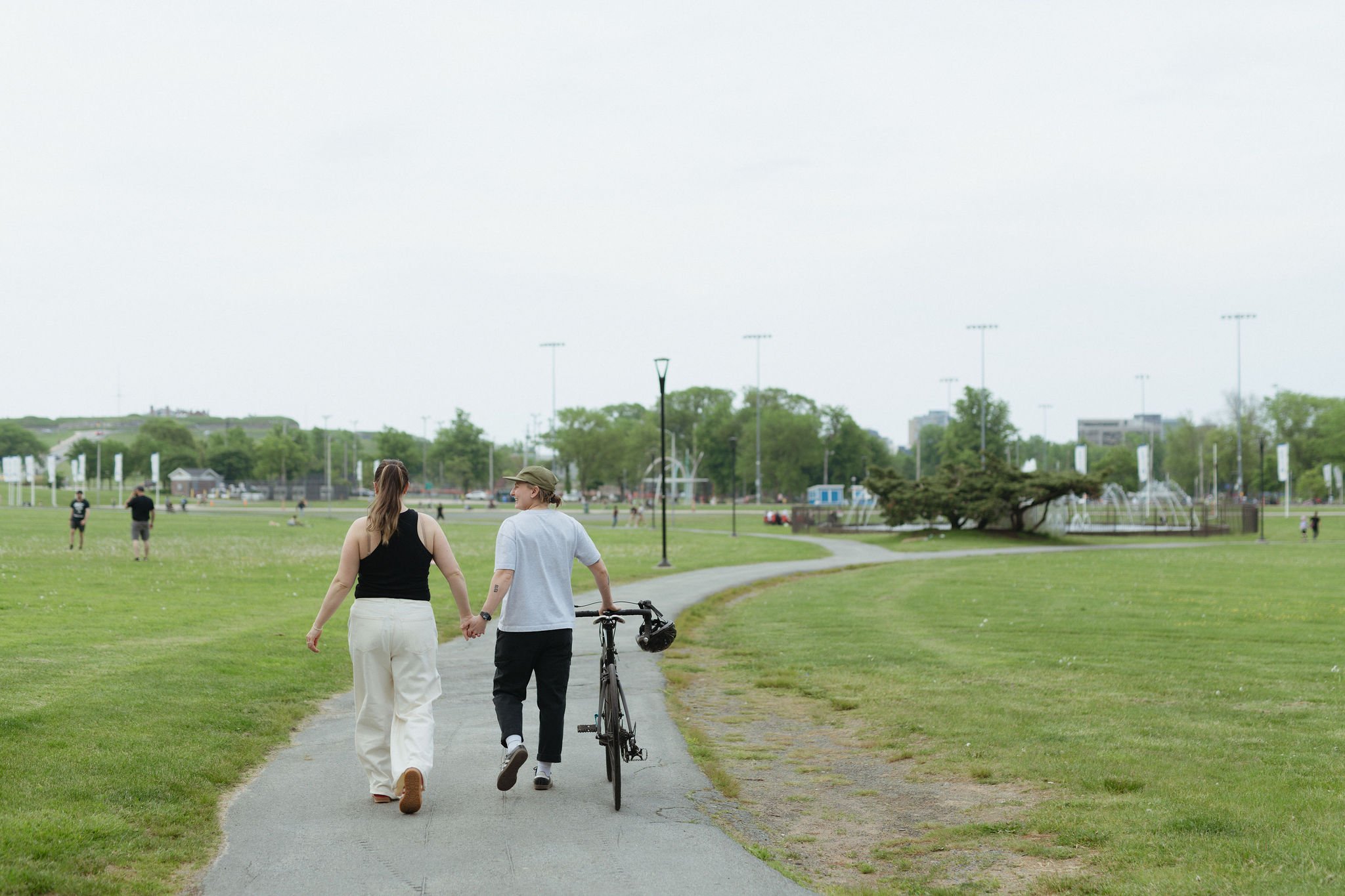 Two people walking hand in hand with a bicycle on a paved pathway in a park with green grass and trees, other park visitors, and distant city buildings in the background.