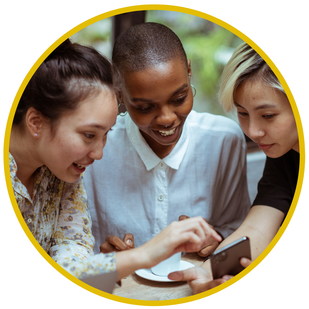 Three diverse women sitting at a table together, looking at a smartphone and smiling.
