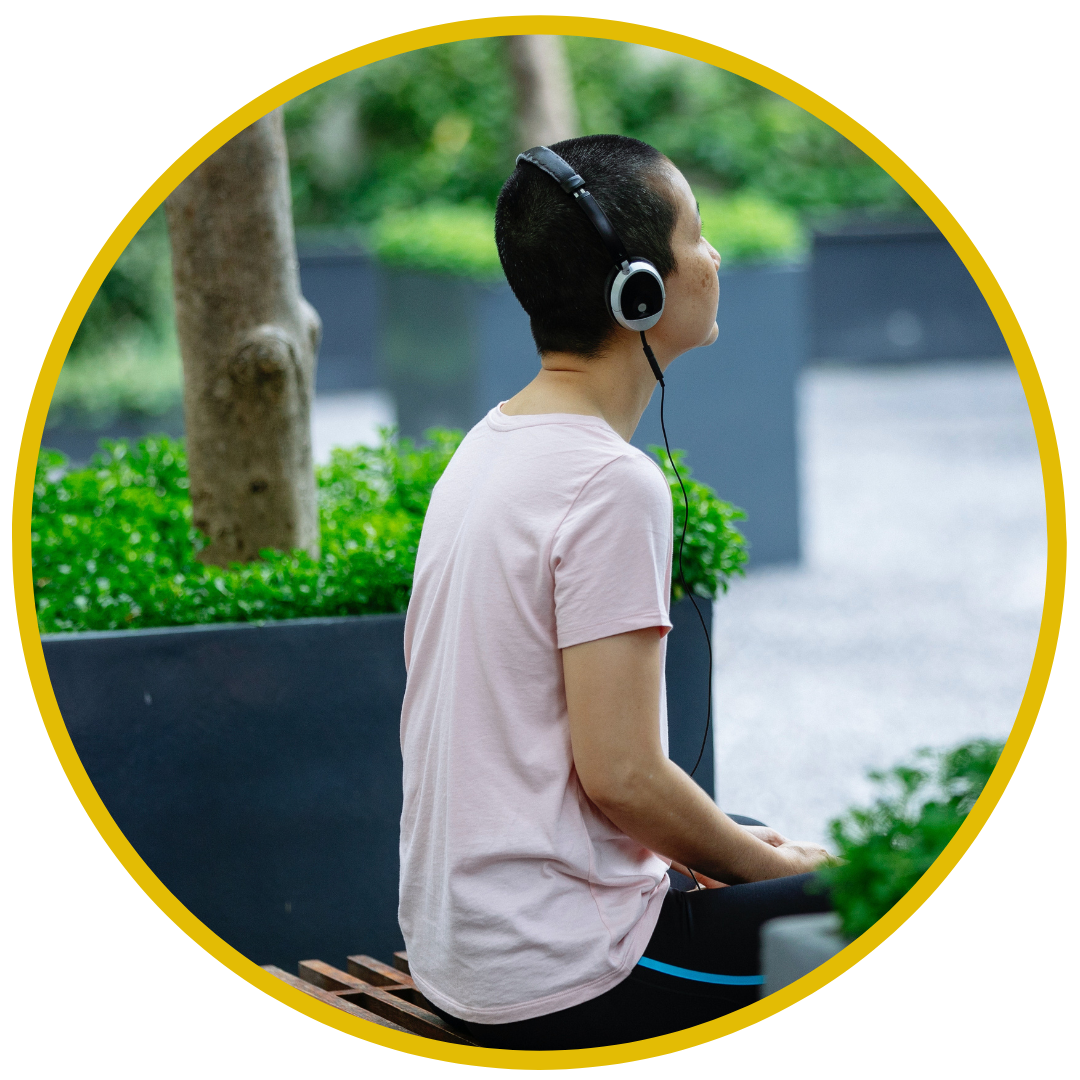 A person with short dark hair sitting on a park bench wearing headphones, looking to the right, with green bushes and trees in the background.