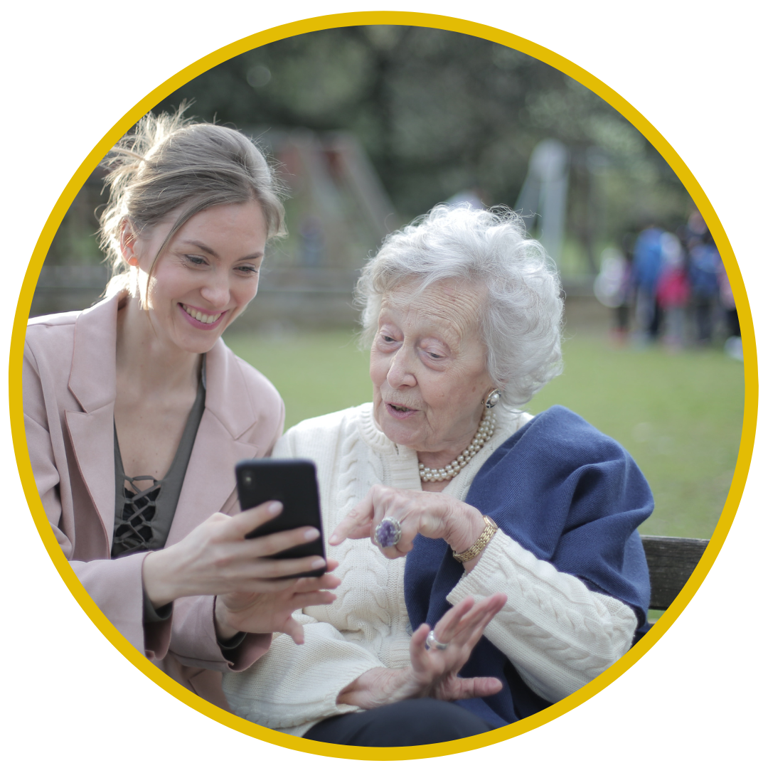 A young woman and an elderly woman are sitting on a park bench, looking at a smartphone. They are smiling and appear to be enjoying their time outdoors.