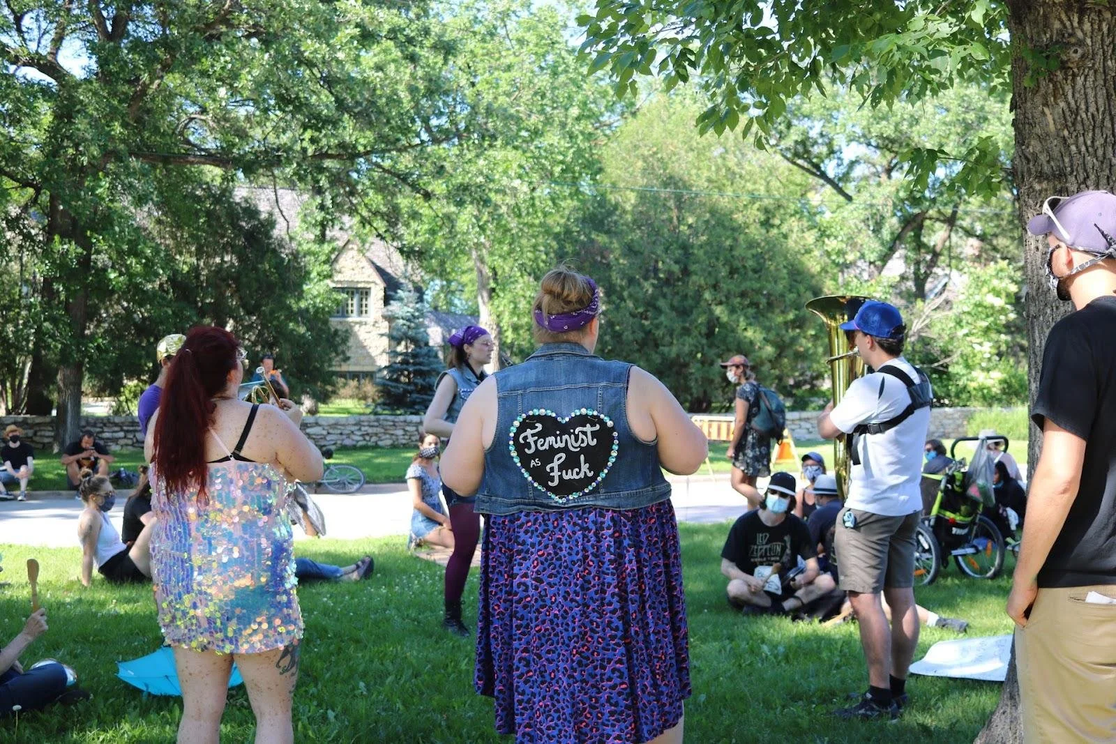 A group of people are relaxing in a park, one person has a vest that says 'feminist as fuck' on it