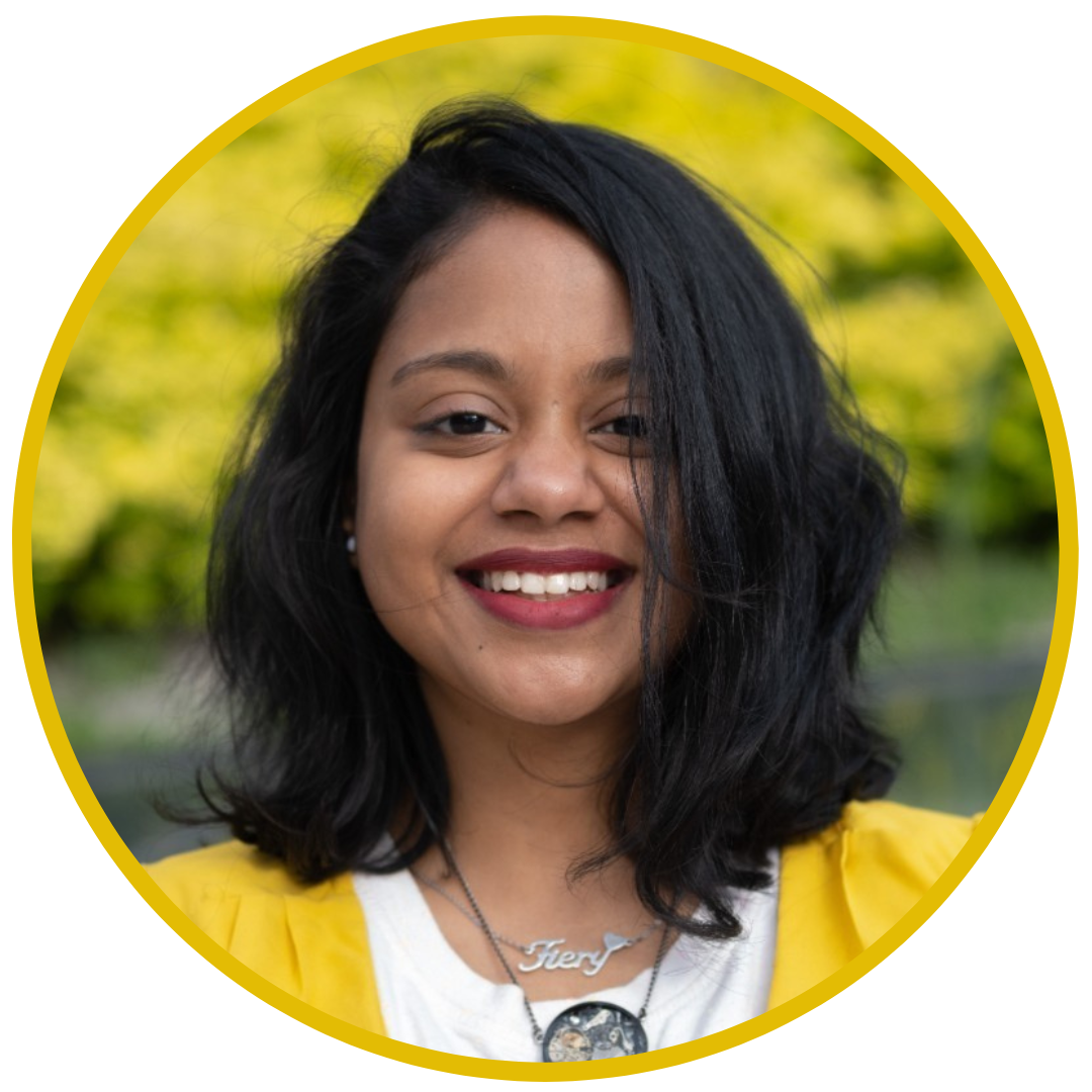 A smiling woman with black hair, wearing a yellow top and necklaces, with a blurred outdoor background.
