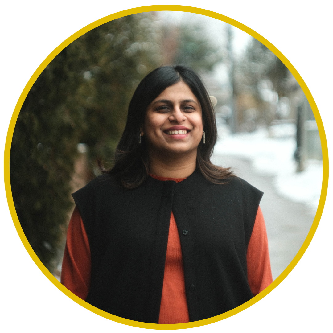 A smiling woman with shoulder-length dark hair, wearing a black vest over an orange shirt, standing outdoors on a winter day with snow on the ground and blurred trees and houses in the background.