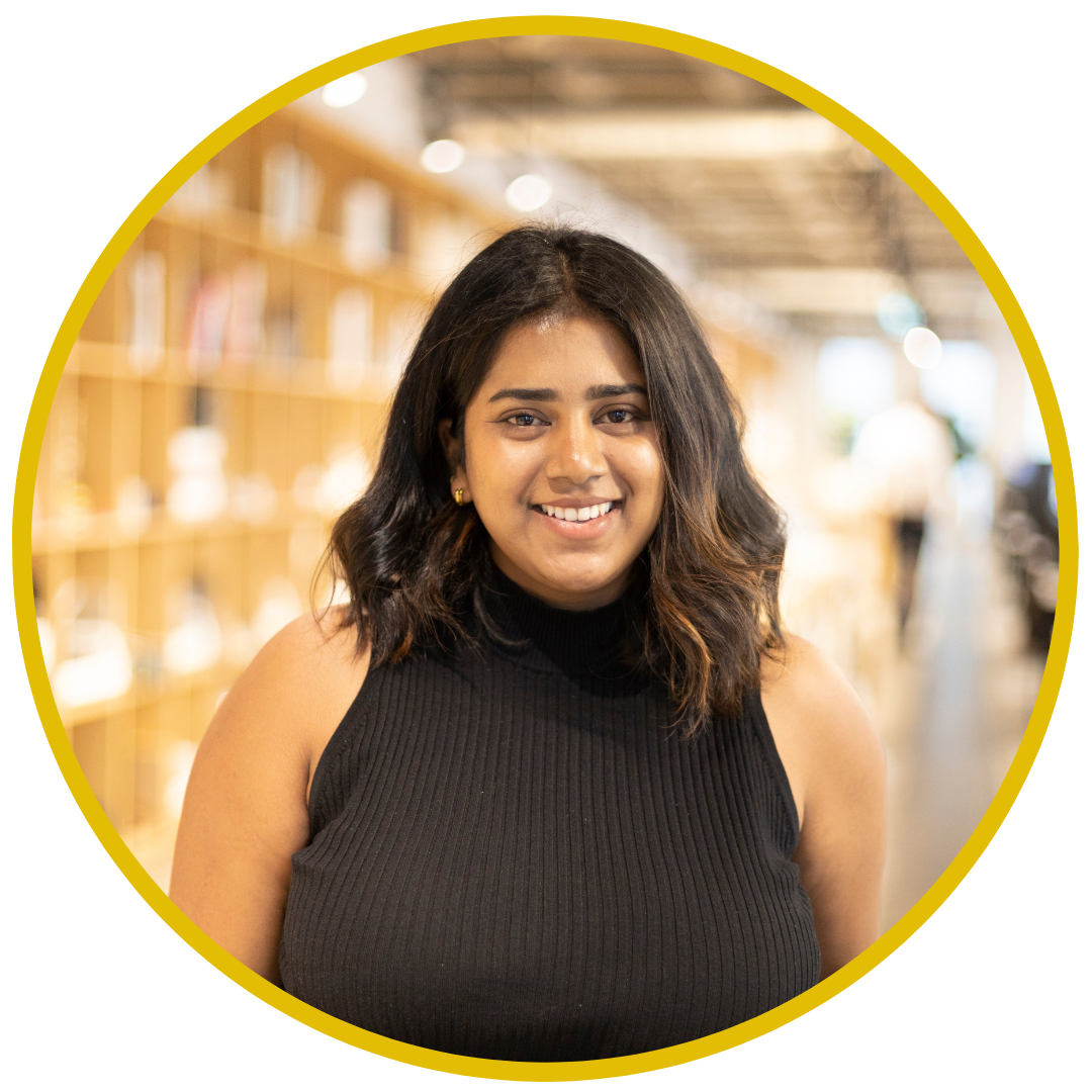A young woman smiling with shoulder-length dark hair, wearing a sleeveless black top, standing in an indoor space with wooden shelves and books.