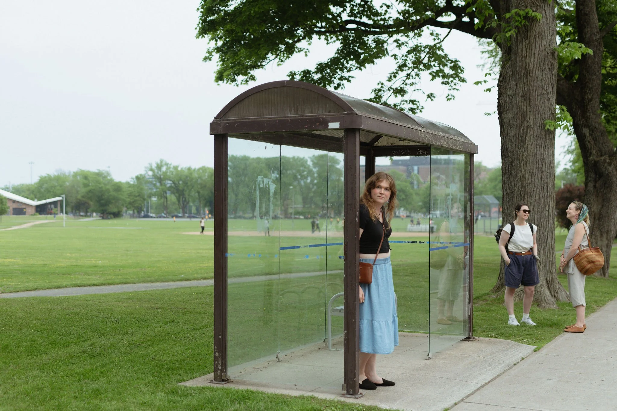A woman standing inside a transparent bus shelter with trees and grass in the park, while two women stand and chat nearby.