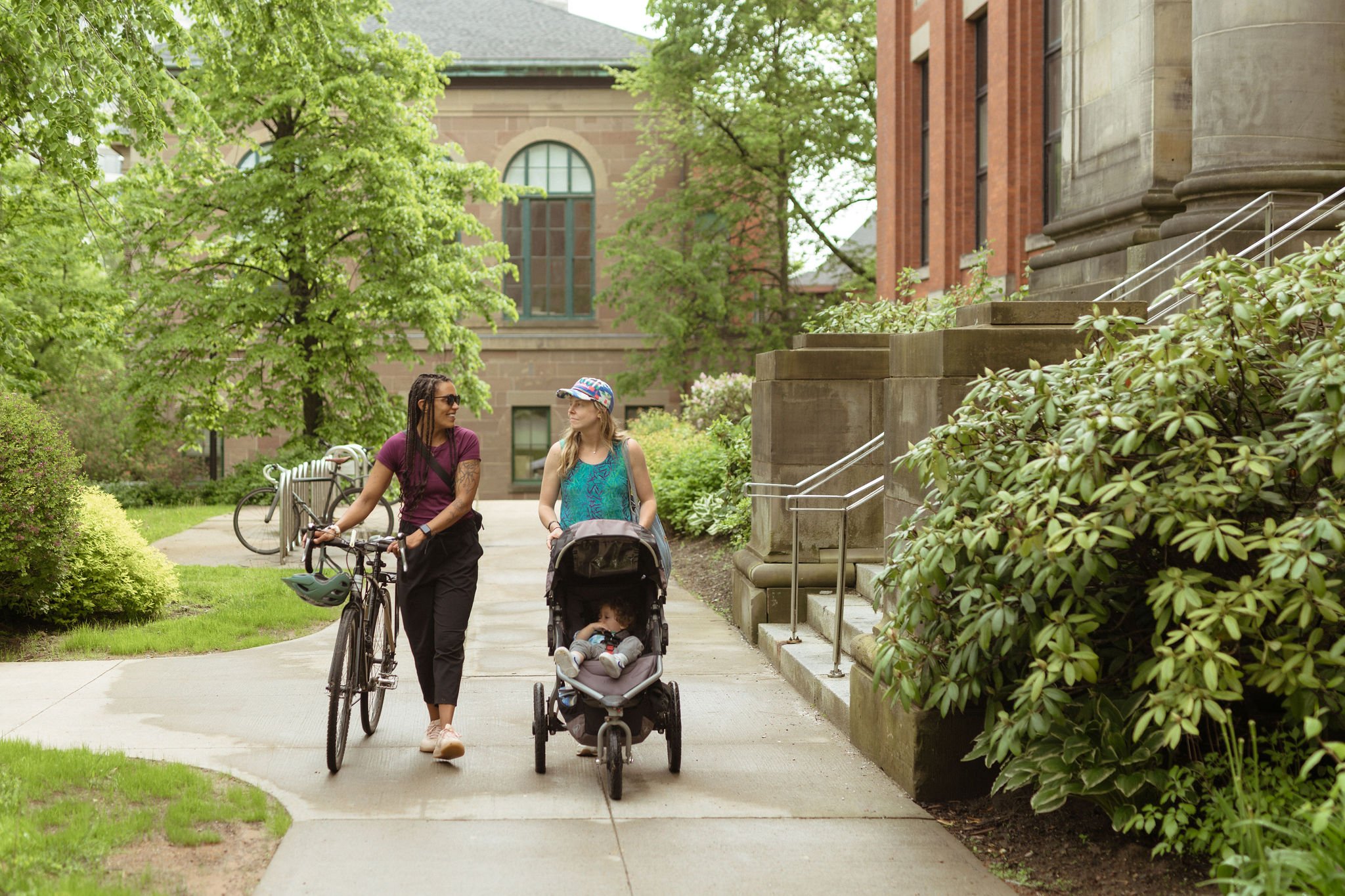 Two women walking on a sidewalk with a bicycle and a stroller, surrounded by greenery and historic buildings.