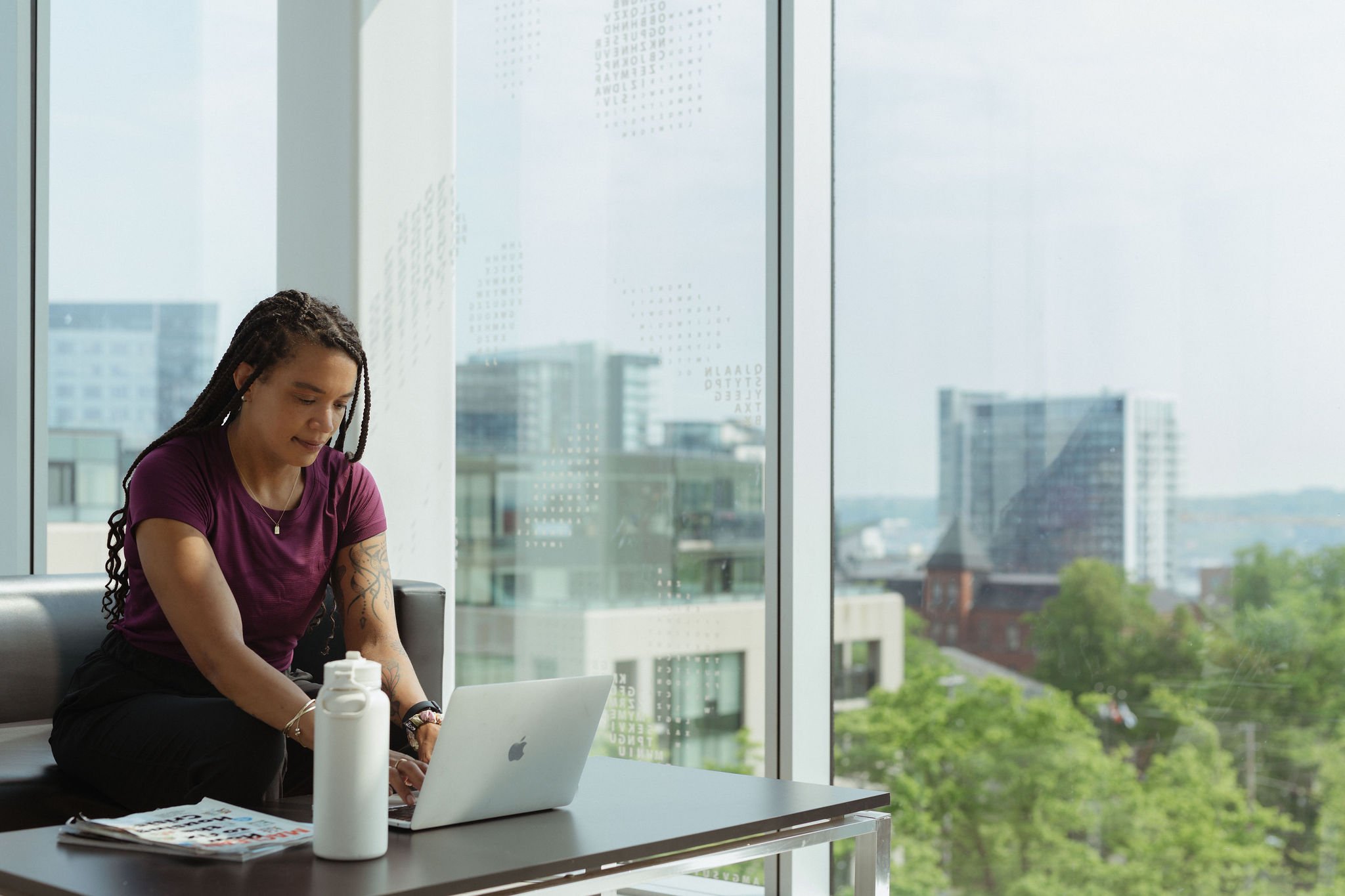 A woman with dreadlocks working on a laptop in a modern office with large windows, city buildings, and green trees outside.