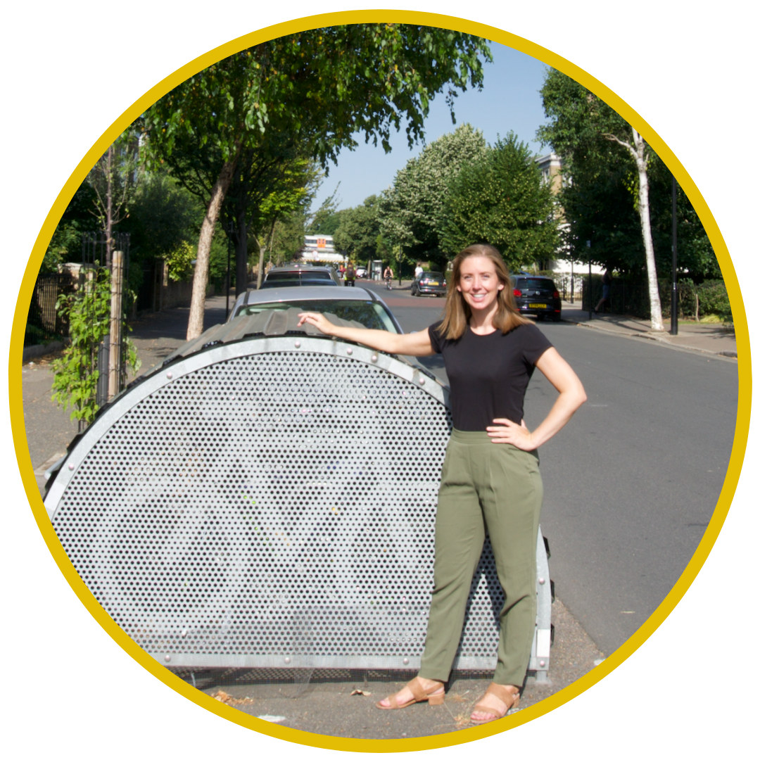 A woman standing next to a large, metallic, curved object on the sidewalk in a suburban neighborhood lined with trees and parked cars under a clear blue sky.
