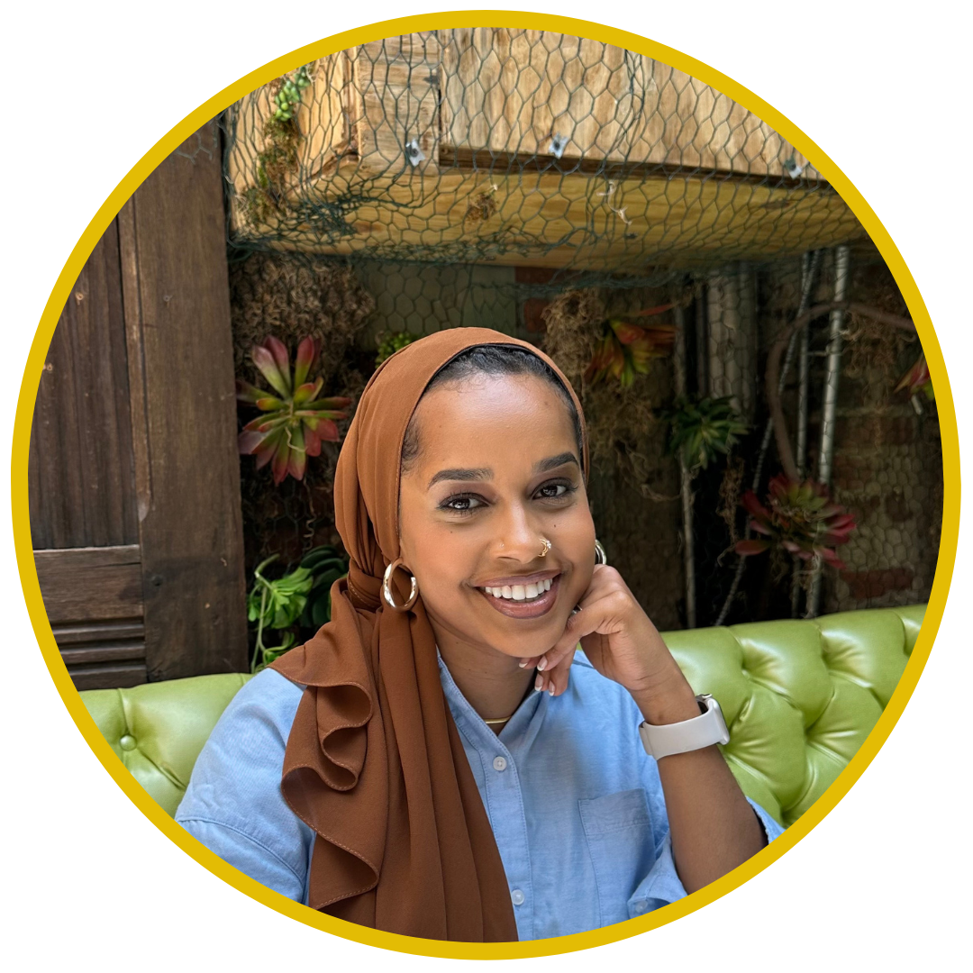 Smiling woman with hoop earrings and a nose ring sitting on a green cushioned seat, wearing a light blue shirt and brown headscarf, in a cozy outdoor setting with plants and wood decor.
