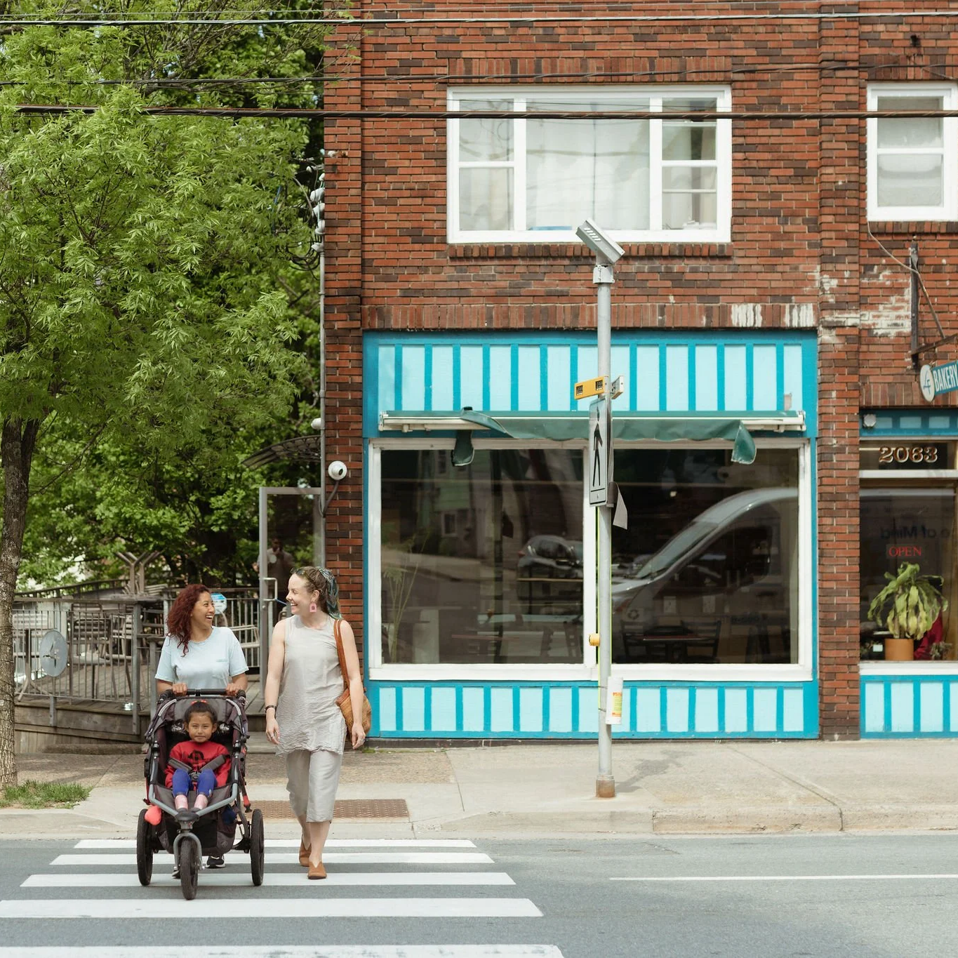 Two women walking across the crosswalk on a city street, one pushing a stroller with a young girl inside, in front of a brick building with large windows and blue trim.