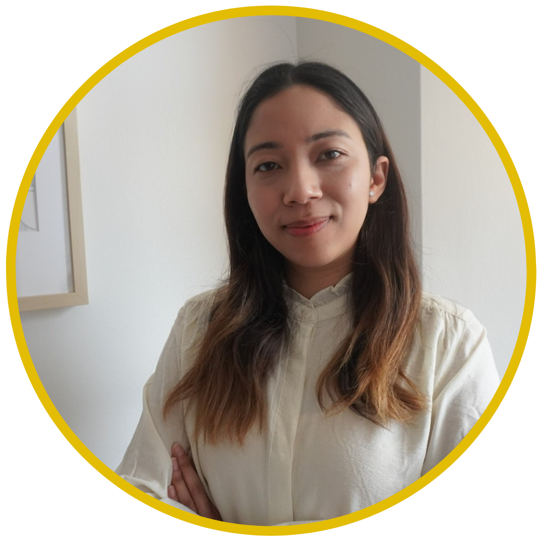A woman with long brown hair, wearing a cream-colored shirt, standing indoors with arms crossed, smiling gently, in front of a neutral background with a white wall and a framed picture.