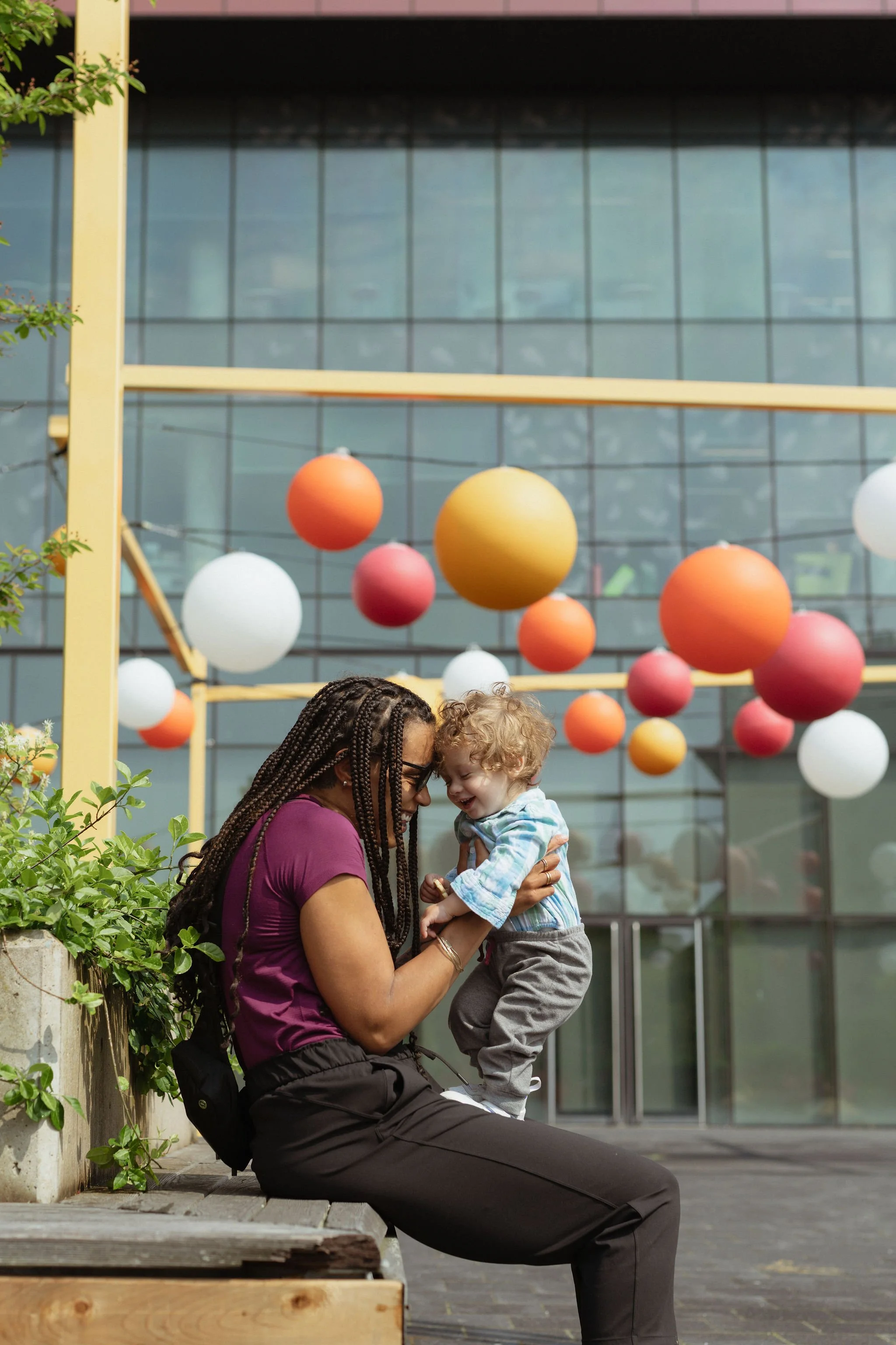 A mother is sitting and holding her child facing her outside a glass building with a bright art installation behind them.