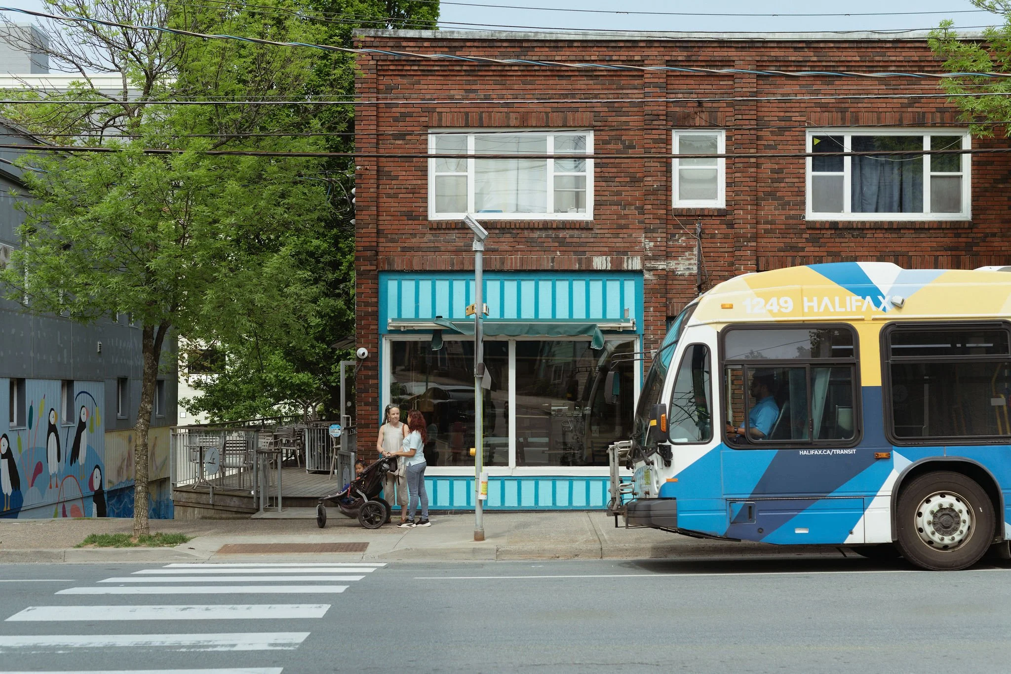 A city sidewalk scene featuring a parked city bus, two women talking with a stroller, and a brick building with large windows. The street has a pedestrian crosswalk and trees nearby.