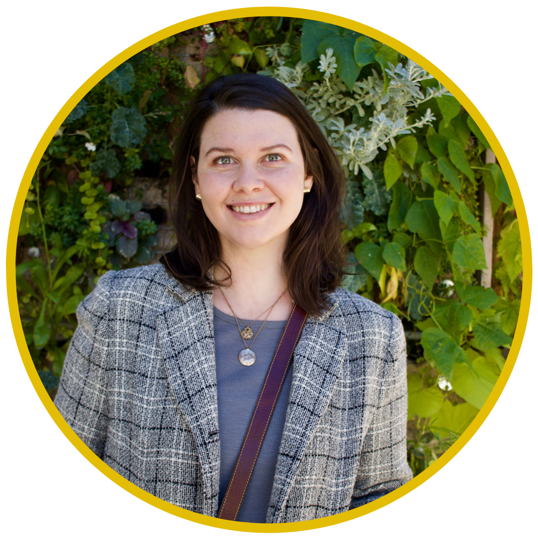 A woman with dark brown hair, wearing a gray blazer and layered necklaces, standing in front of a green leafy background.