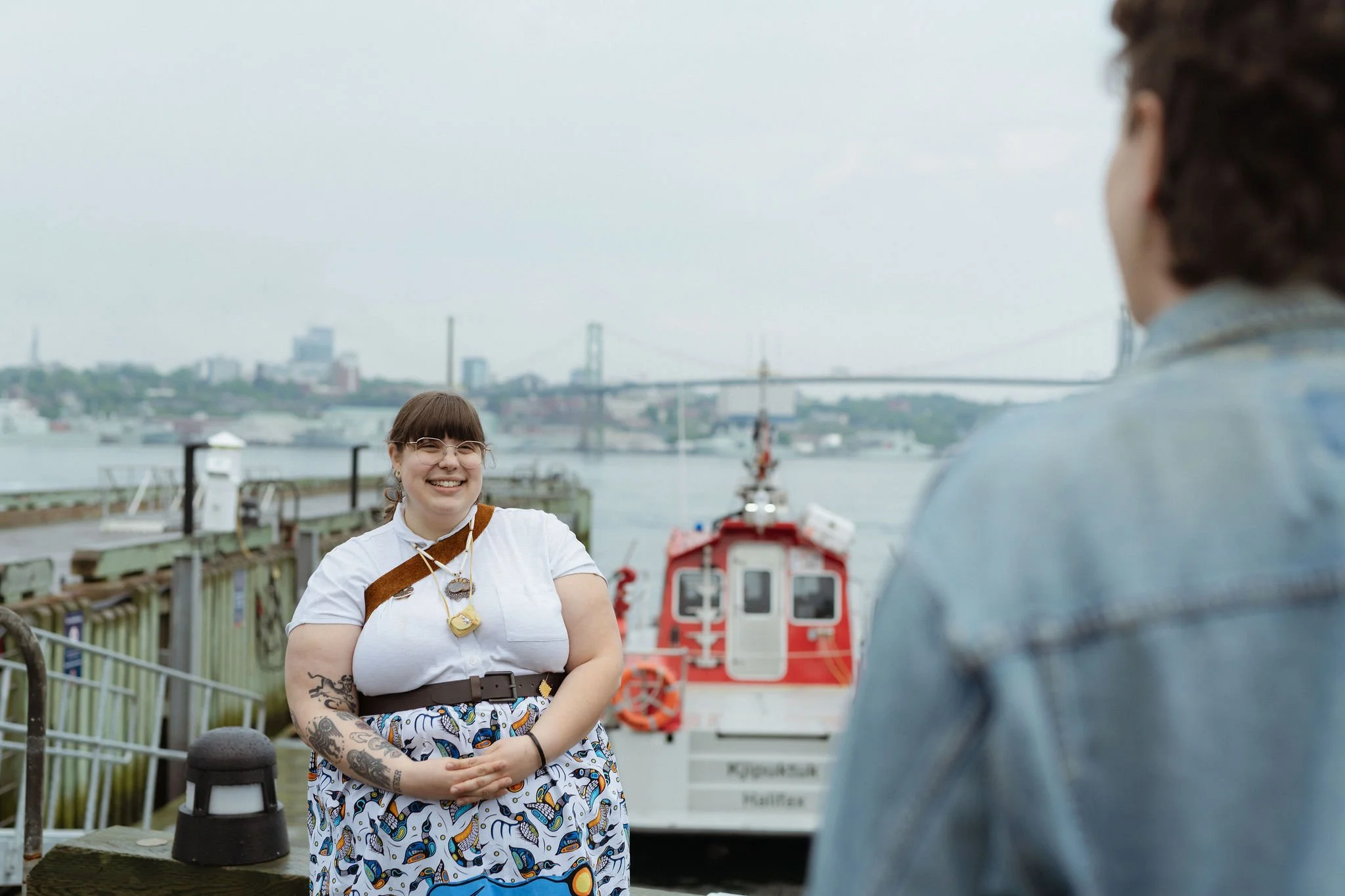A woman with tattoos wearing glasses, a white shirt, and a patterned skirt, smiling and talking to another person by the water dock with boats in the background and a bridge in the distance.