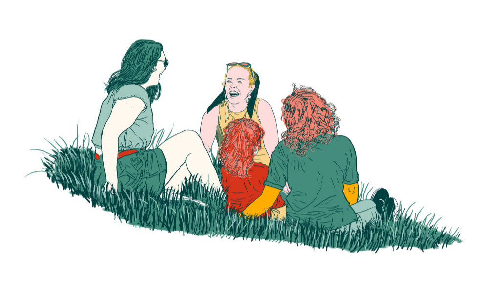 Four young women sitting on the grass in front of a green chalkboard, engaged in conversation and laughing.