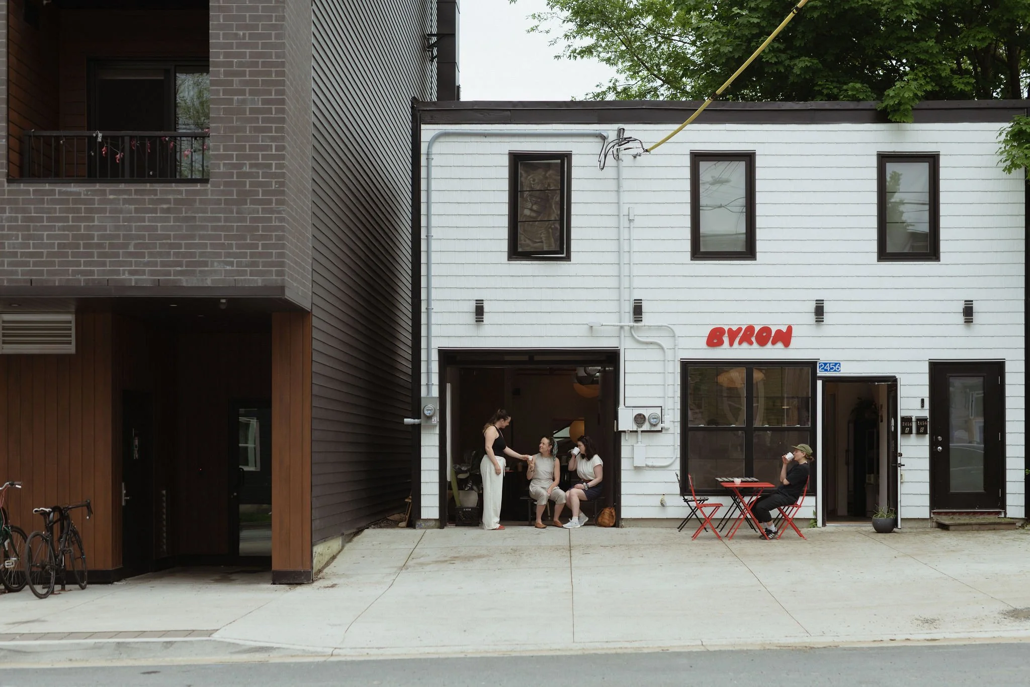Three women sitting and chatting outside a cafe named Byron, with one woman standing and handing something to two women sitting on a bench, and another woman sitting at a red table drinking from a cup, in front of a white building with black-framed windows.