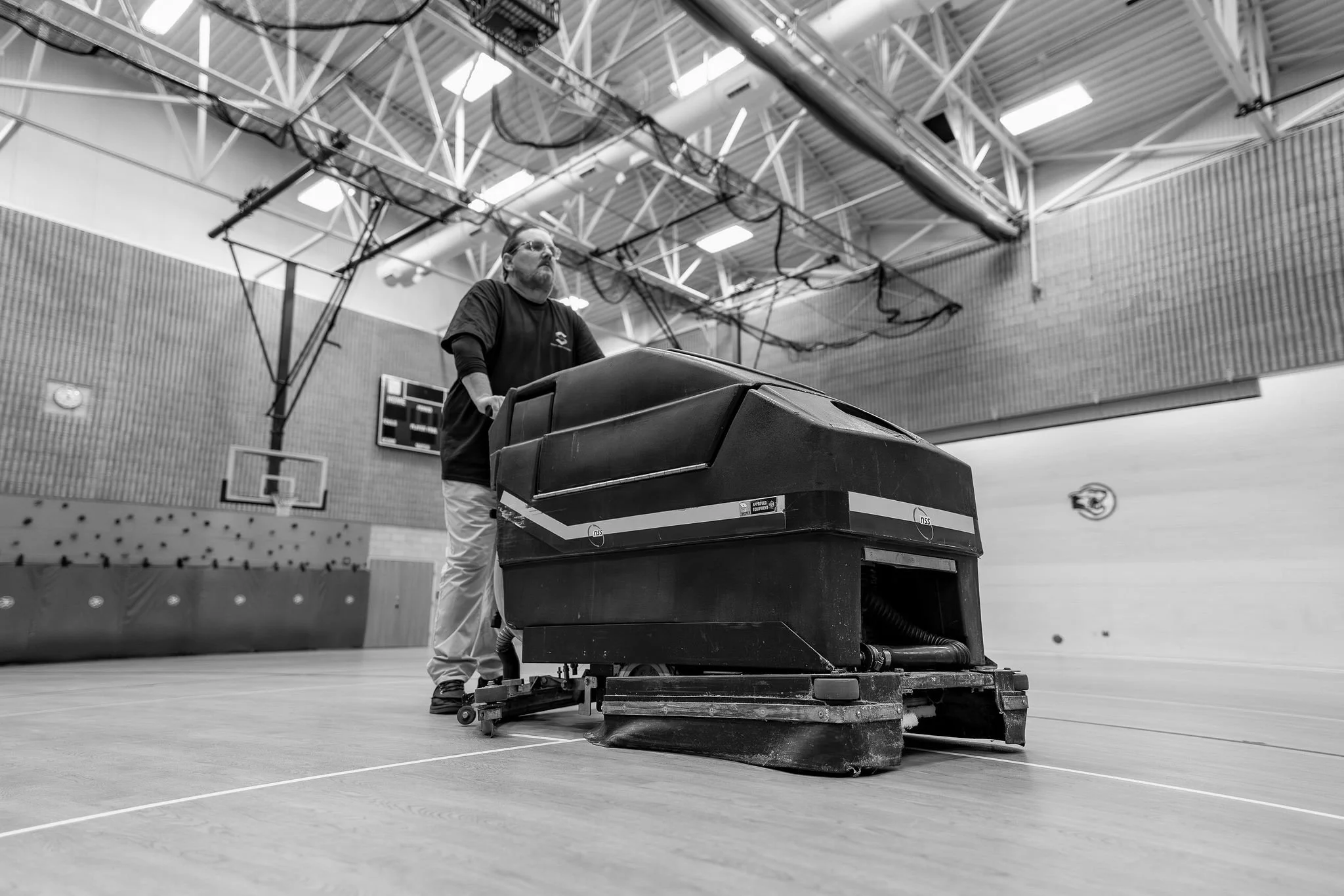 A person operating a floor cleaning machine inside a gymnasium with basketball hoops and climbing equipment in the background.