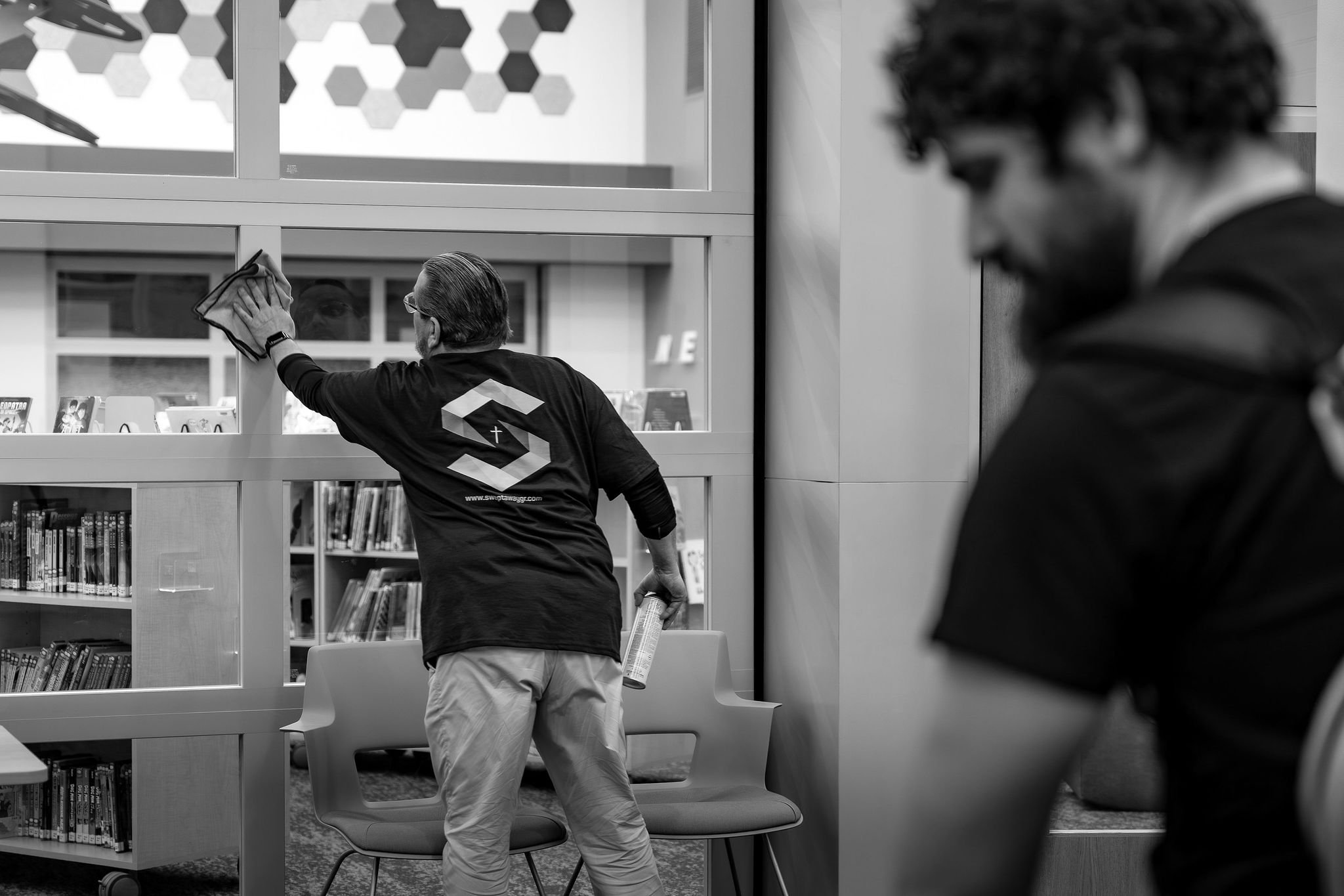 Two men cleaning windows in a library or bookstore, one is cleaning higher on the window with a cloth, and the other is observing or waiting nearby holding a spray can.