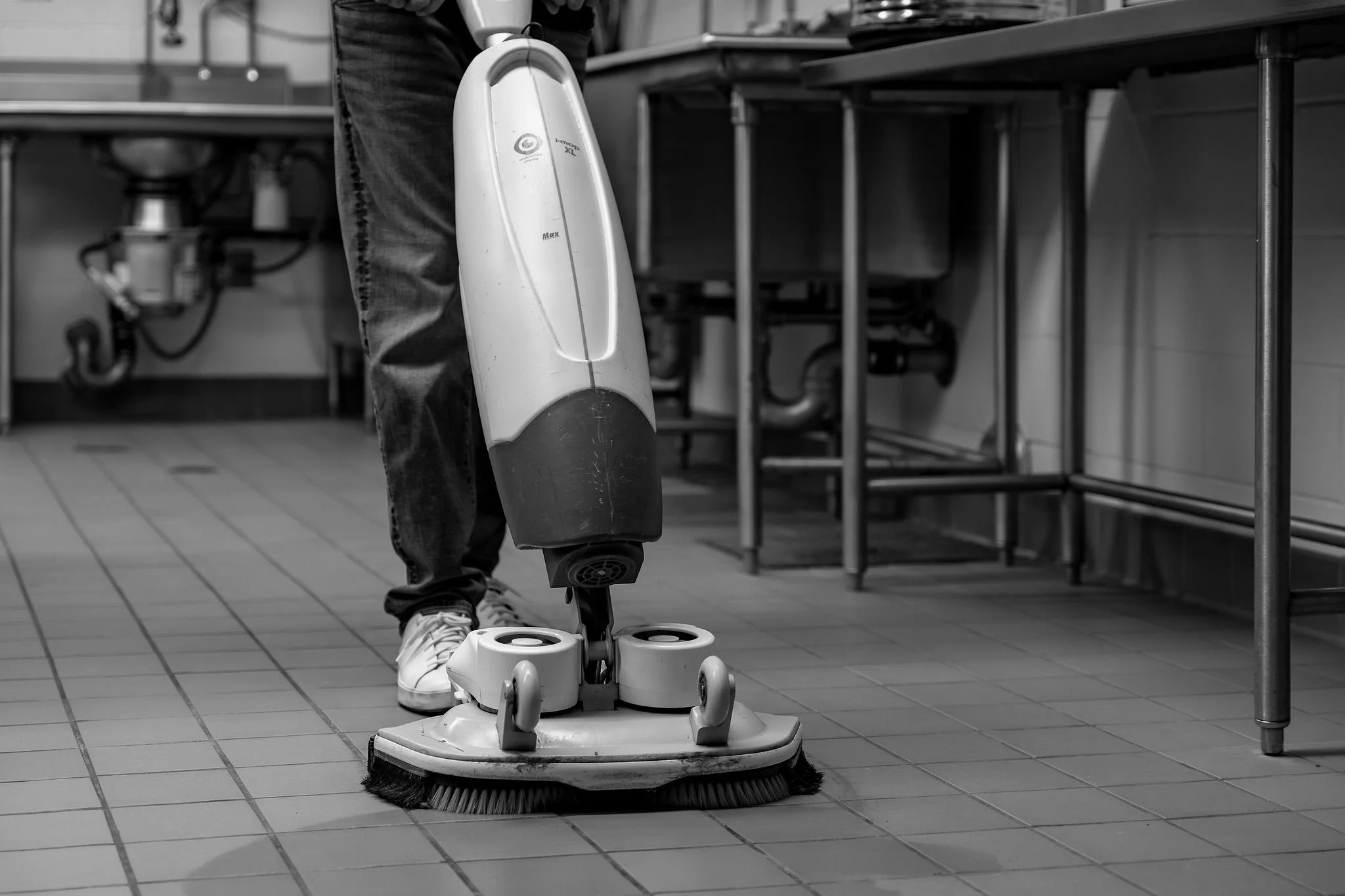 A person using a floor cleaning machine in a commercial kitchen with tiled floor and metal worktables in the background.