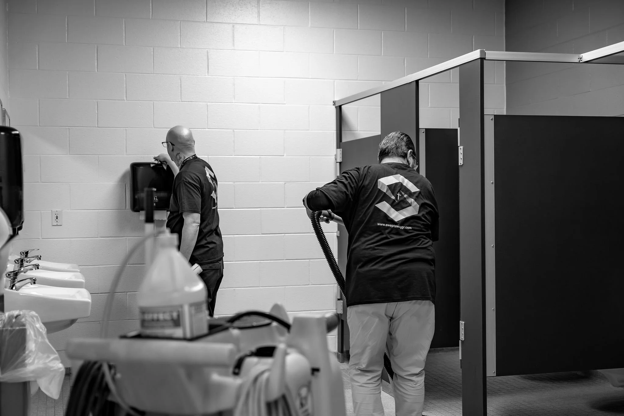 Two men working in a restroom, one at a sink and the other cleaning a bathroom stall.