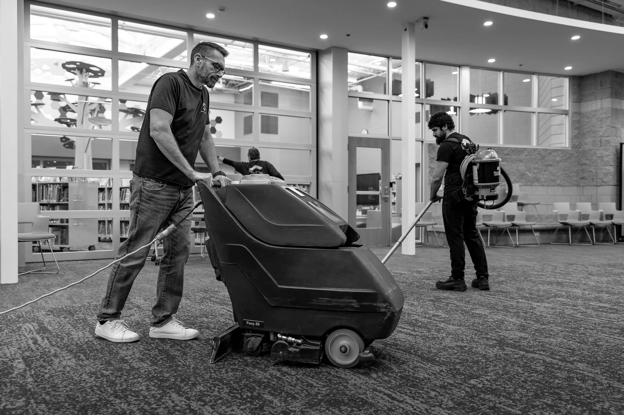 Two workers cleaning a large indoor space, one operating a floor cleaning machine and the other using a floor vacuum, inside a modern building with large windows and seating area.