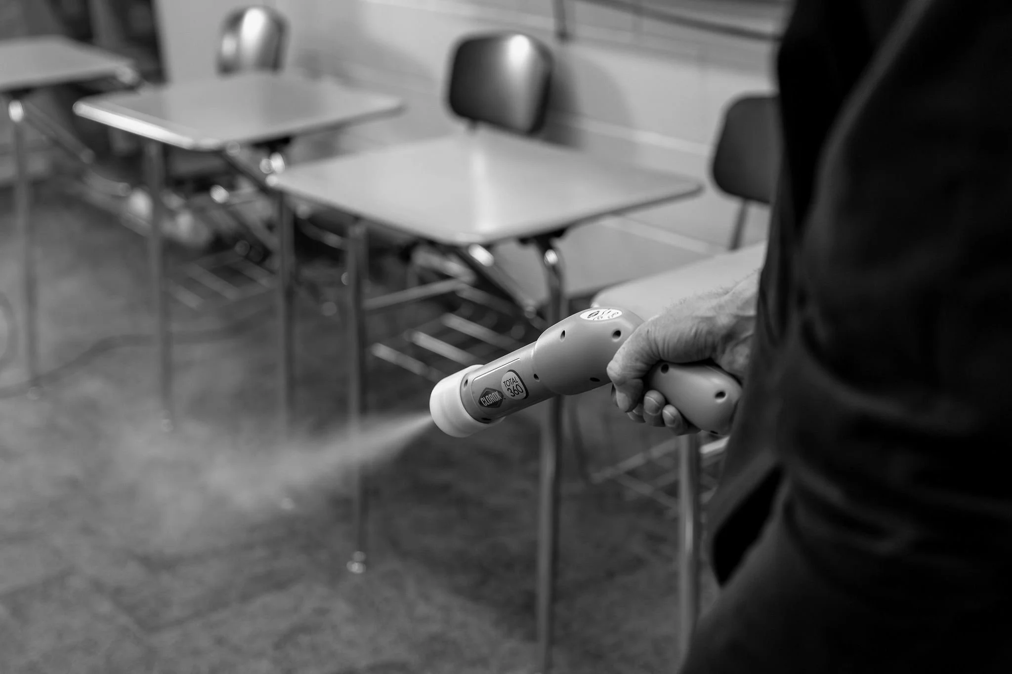 Person disinfecting classroom desks with a handheld spray device in a school room.