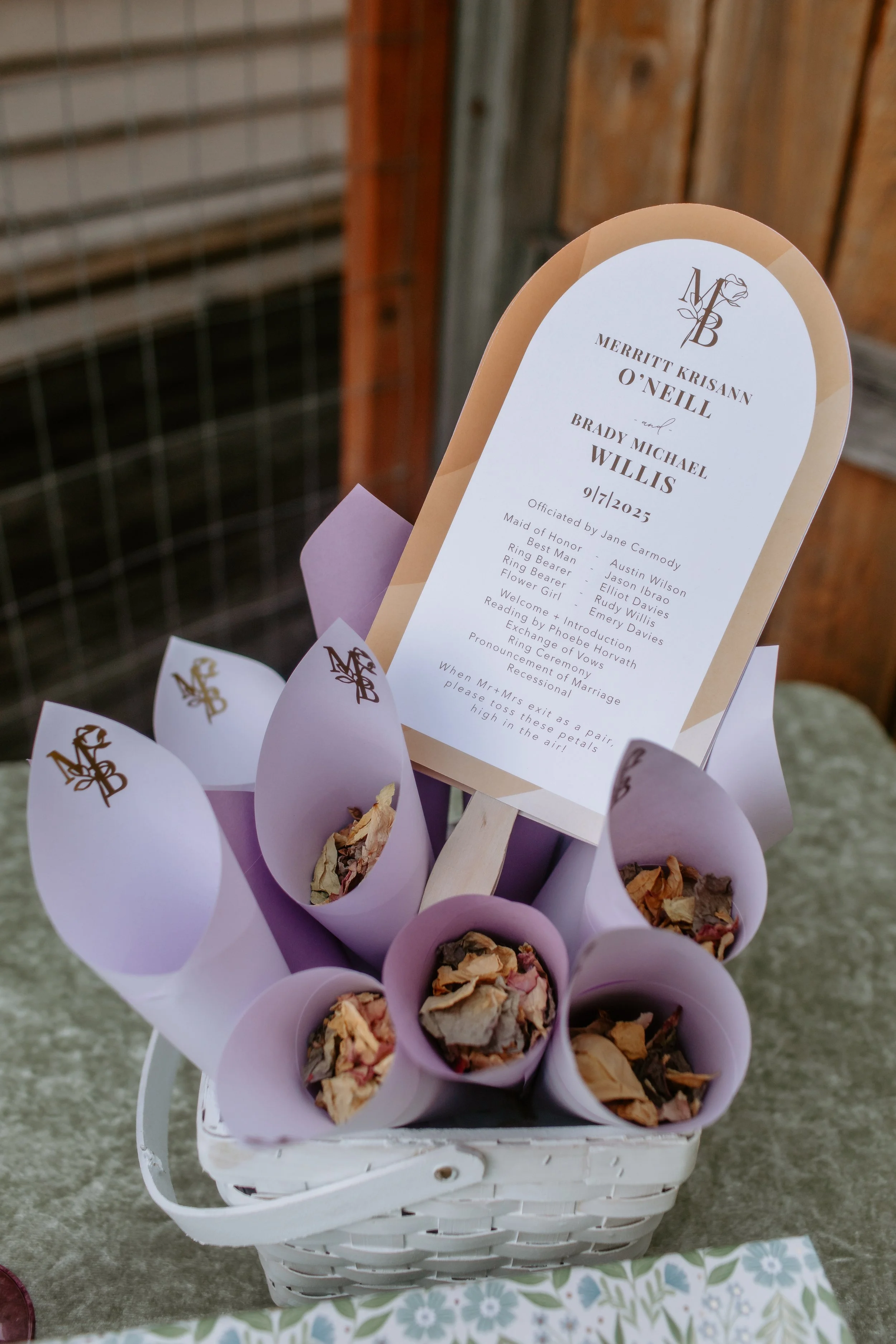 A white basket containing purple paper cones filled with dried flower petals, with a wedding program inserted in the center. The program displays the names Merritt Krisann O'Neill and Brady Michael Willis, and the date 9/17/2025, with details about the wedding ceremony and traditions.