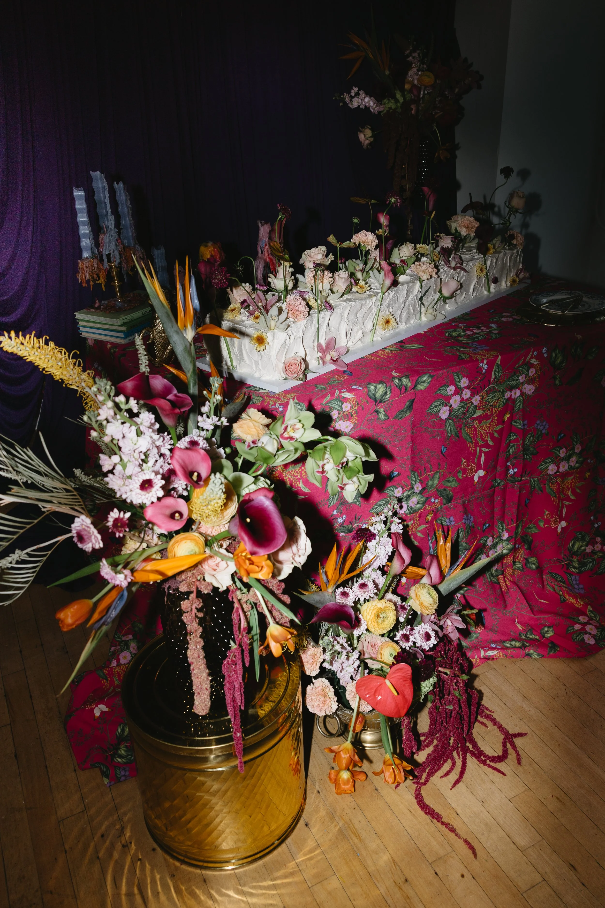 Colorful flower arrangements with exotic flowers in golden vases, set against a purple backdrop and floral tablecloth at a celebration event with decorative candles and books.